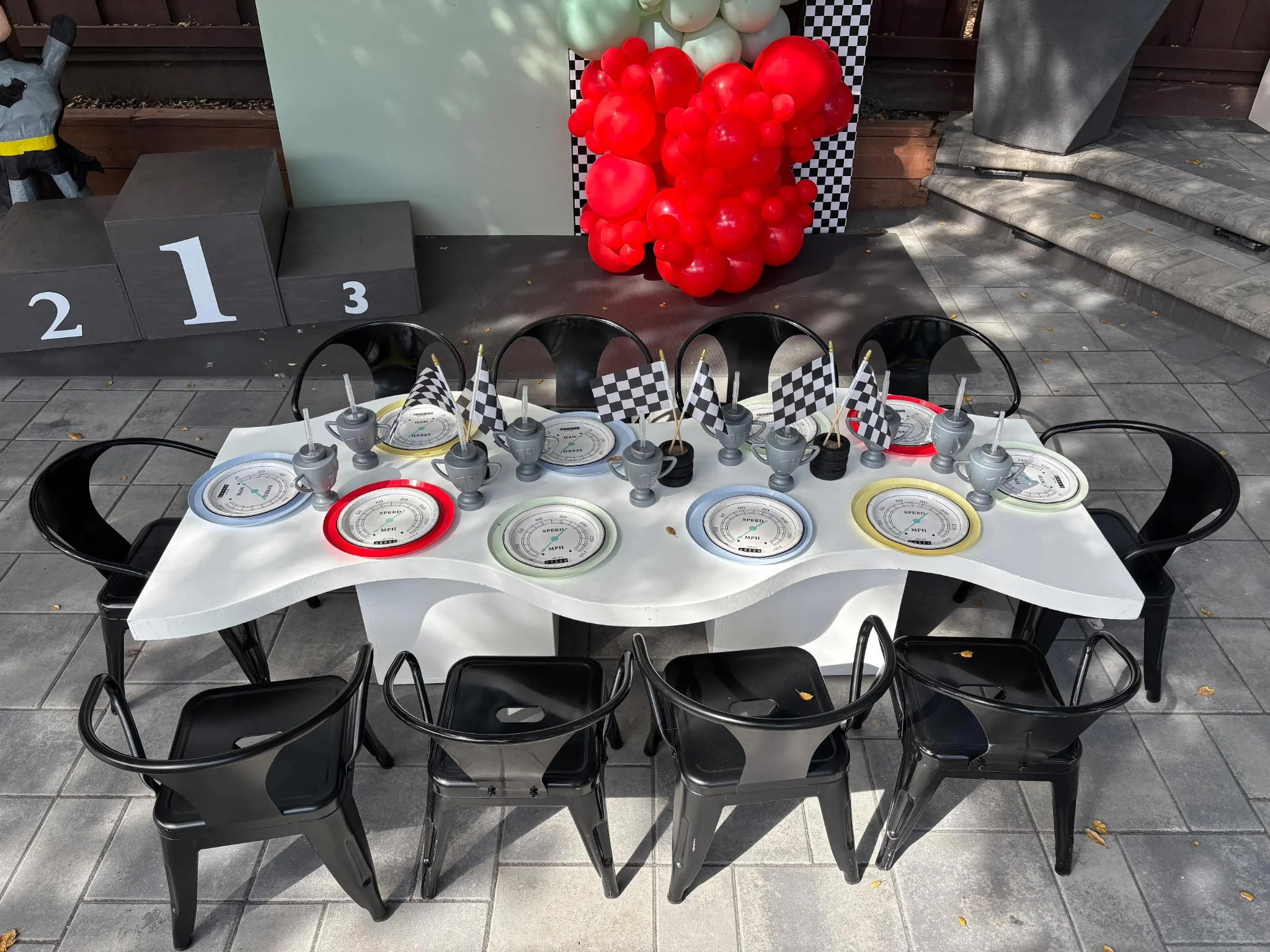 Table with decorations for a racing-themed party, featuring small trophies, checkered flags, and plates, with a backdrop of red and gray balloons and a winners' podium in the background.