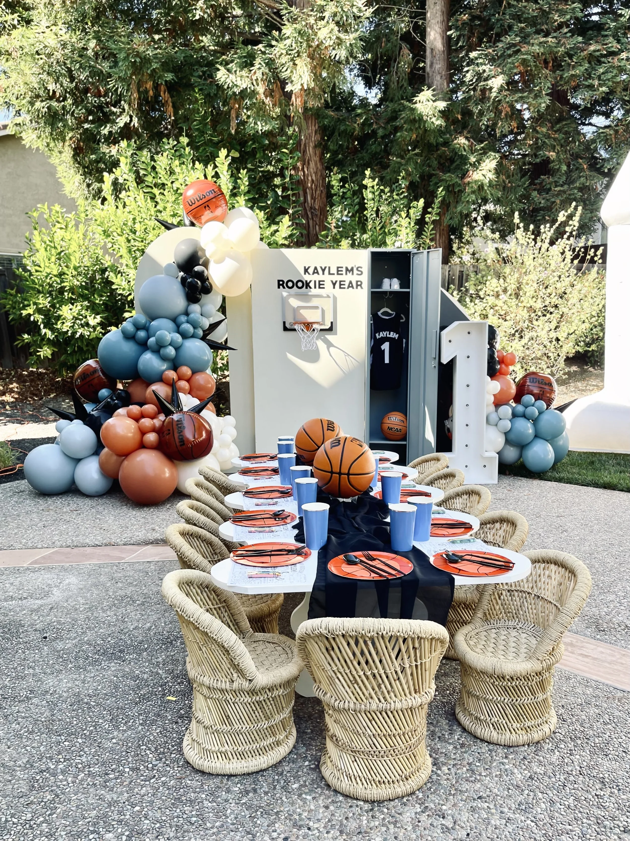 Outdoor celebration setup with a picnic table, navy blue and orange themed decor, balloons, basketball-themed cake, and sports lockers with the text "Kaylem's Rookie Year."