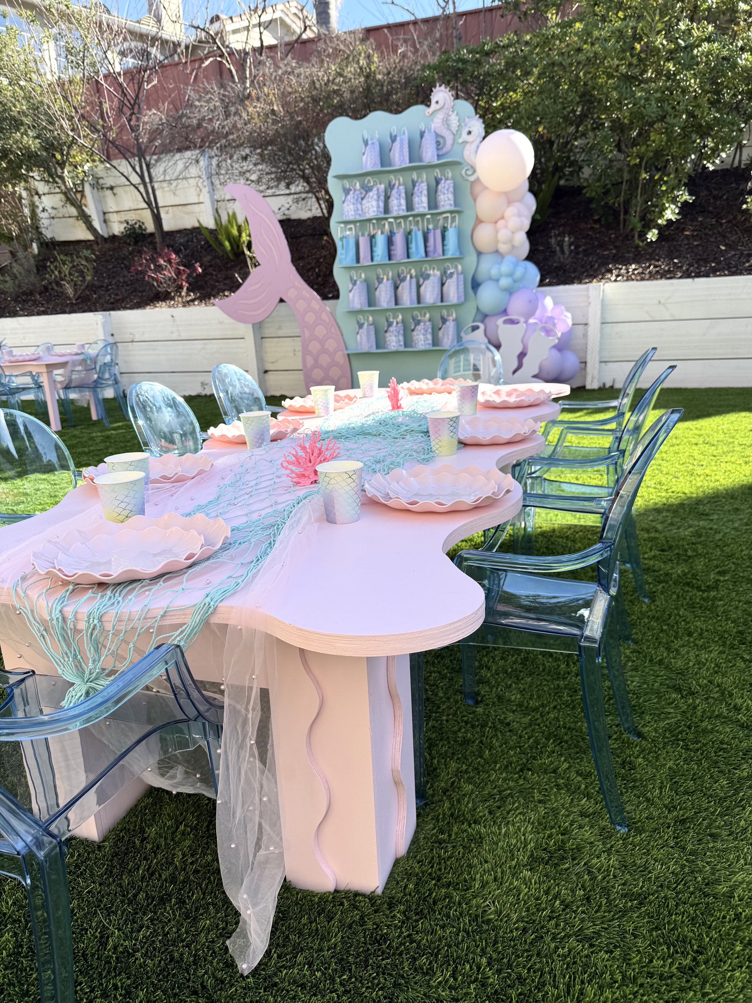 Outdoor party setup with a pink table, clear blue chairs, pastel-colored plates and cups, a decorative pink mermaid tail, and a pastel seahorse-themed backdrop with balloons, all on a grassy backyard.