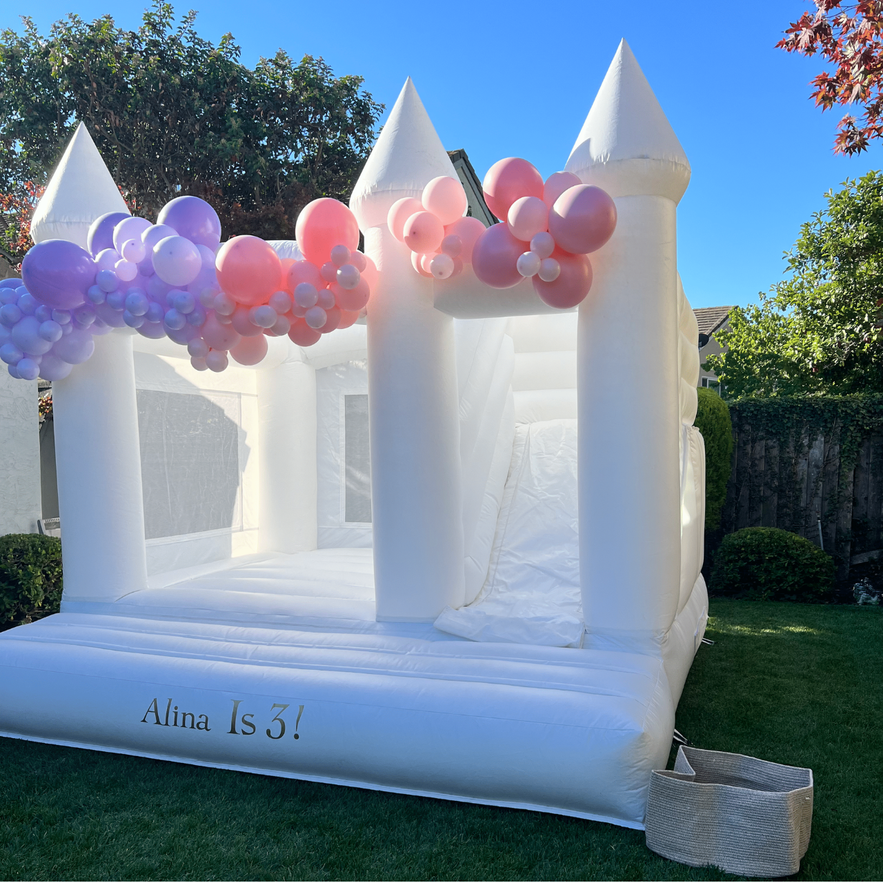 Inflatable white castle bounce house decorated with pink and purple balloons, placed on a lawn with trees and a fence in the background, celebrating Alina's 3rd birthday.