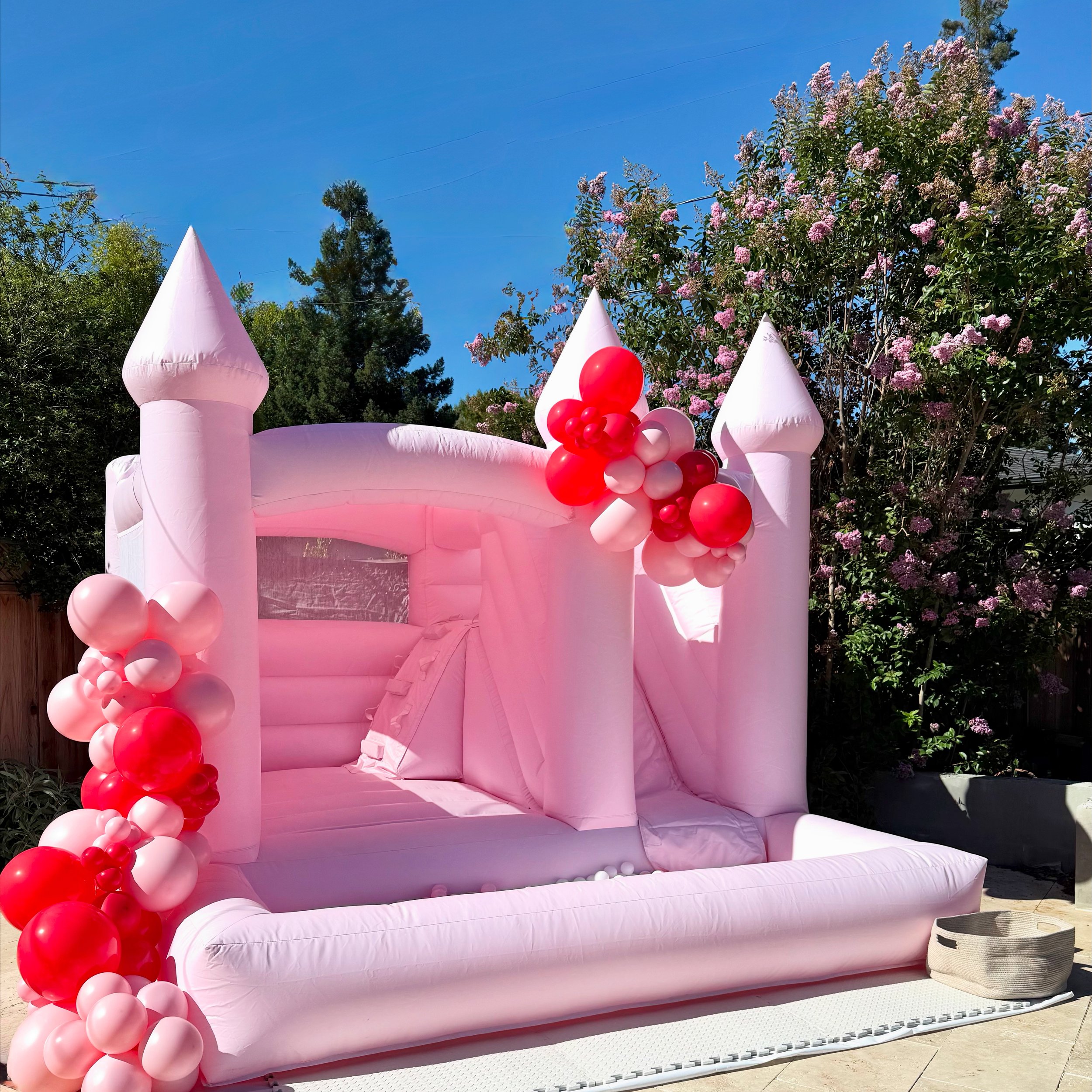 Pink inflatable castle decorated with pink and red balloons, set outdoors with trees in the background and a clear blue sky.