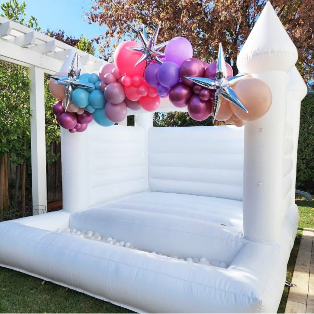 Inflatable photo booth with a balloon garland featuring pink, purple, blue, and peach balloons and silver star-shaped balloons above a white inflatable structure outdoors.