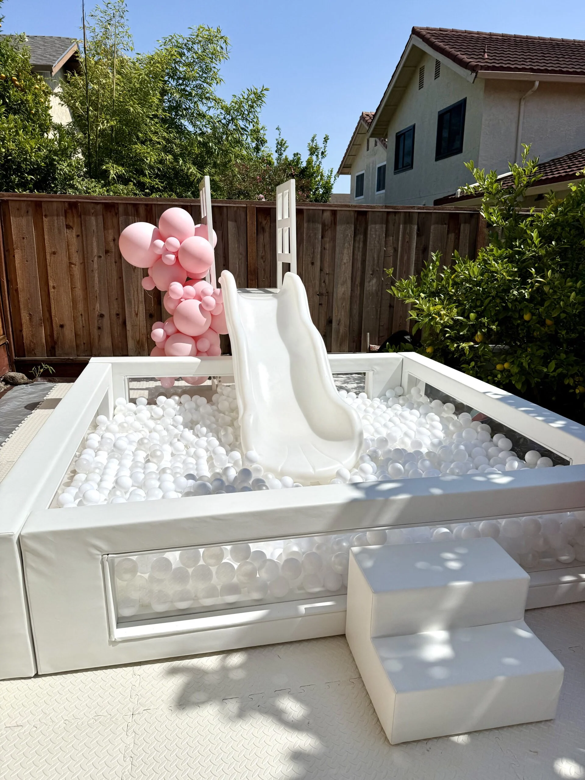 Backyard with a small white ball pit that has a slide and pink balloons attached to a ladder.
