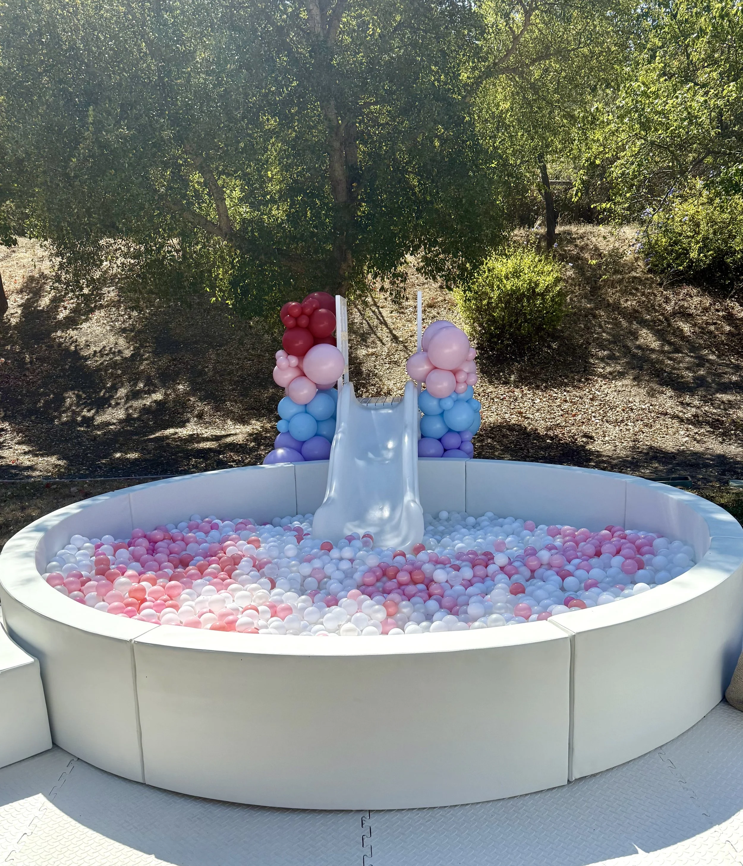 A small ball pit with pink, purple, white, and blue balls, featuring a slide and balloon decorations, outdoors with trees in the background.