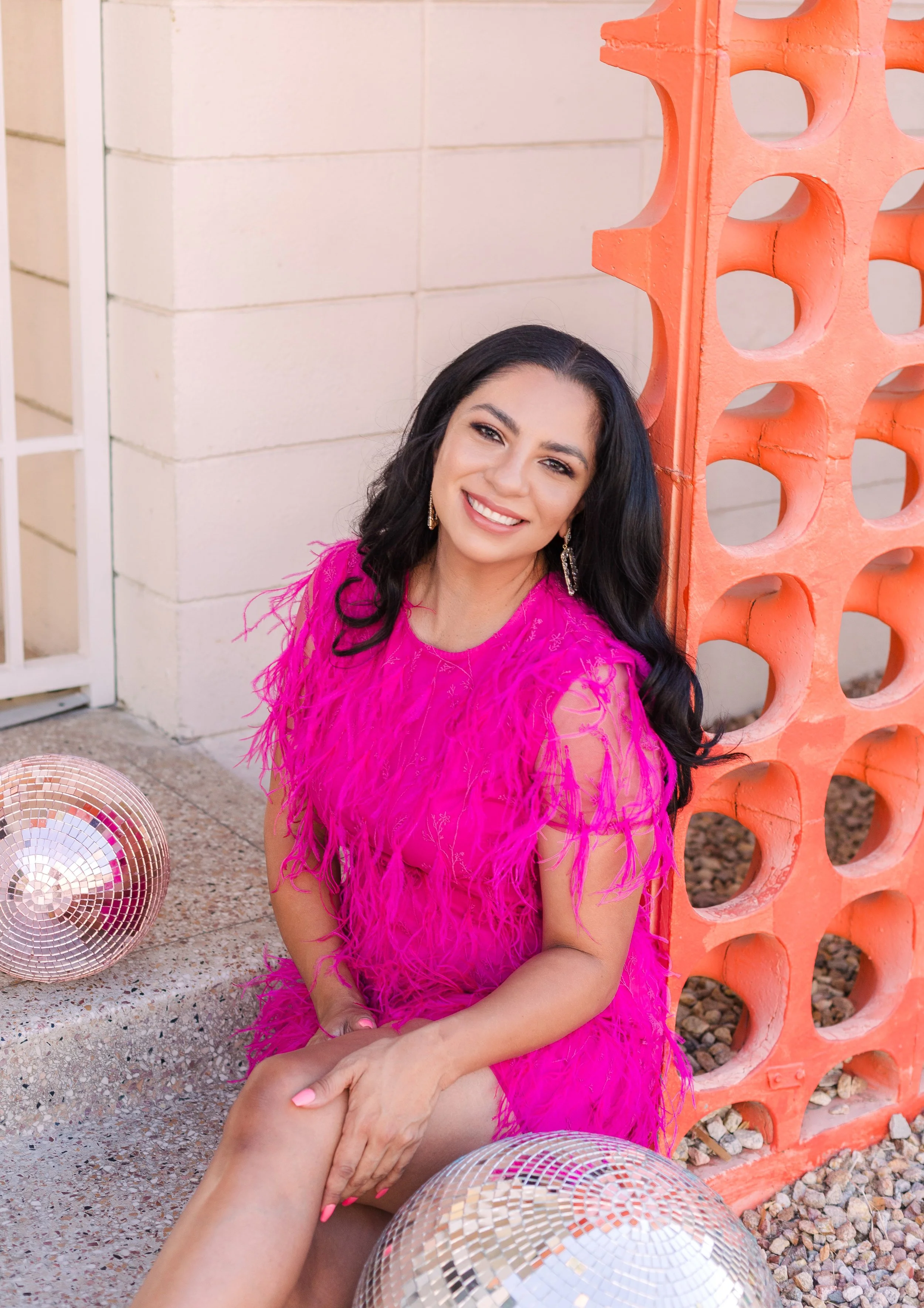 A woman with long black hair, wearing a bright pink feathered dress, is sitting on the ground next to an orange decorative block wall, smiling at the camera. There are two disco balls on the ground, one in front and the other nearby, and a white brick wall is in the background.