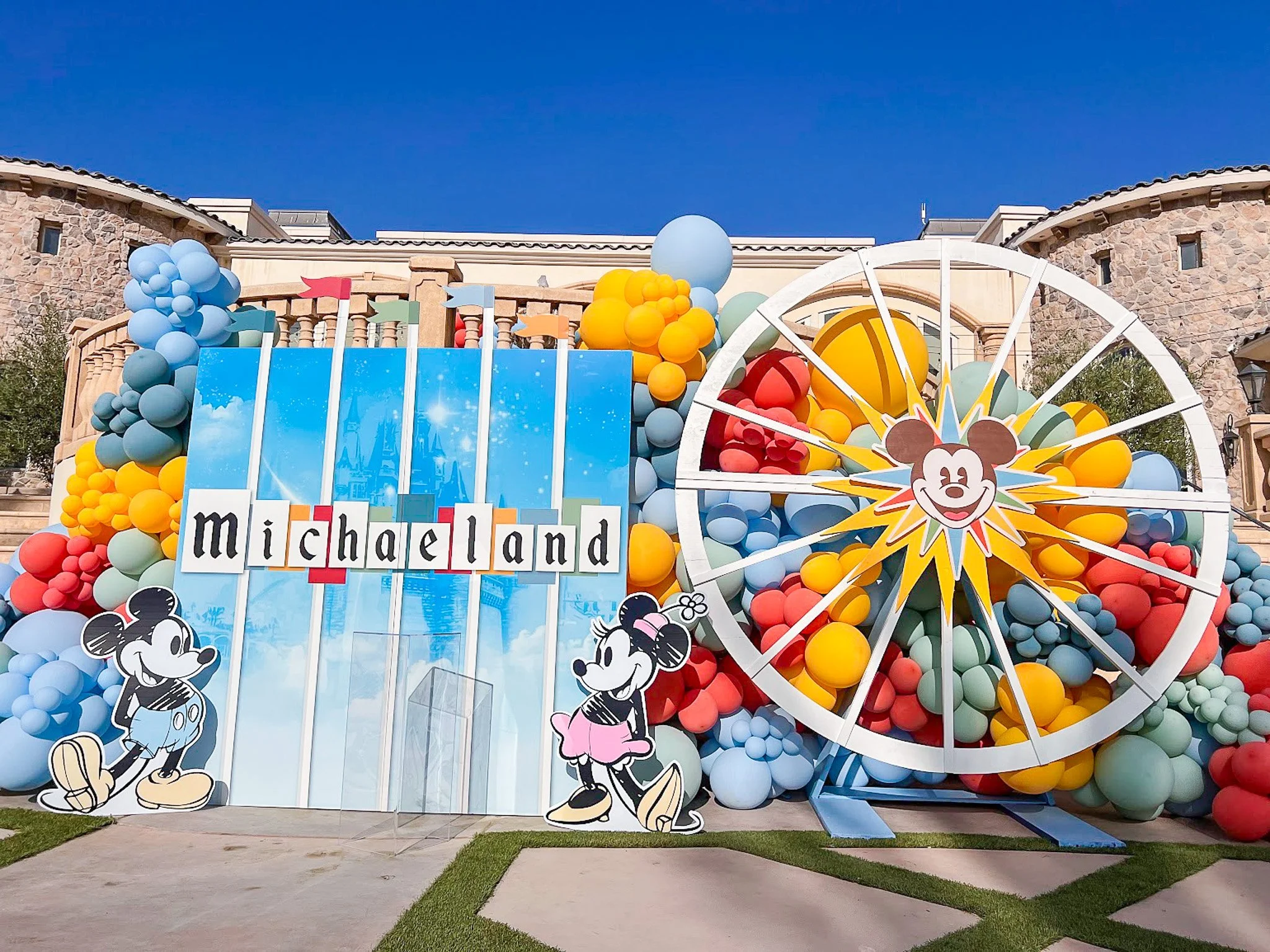 Decorative display with balloons and Disney characters at Mickey's Toontown, featuring Mickey Mouse, Minnie Mouse, and a colorful backdrop with a Ferris wheel and blue sky.