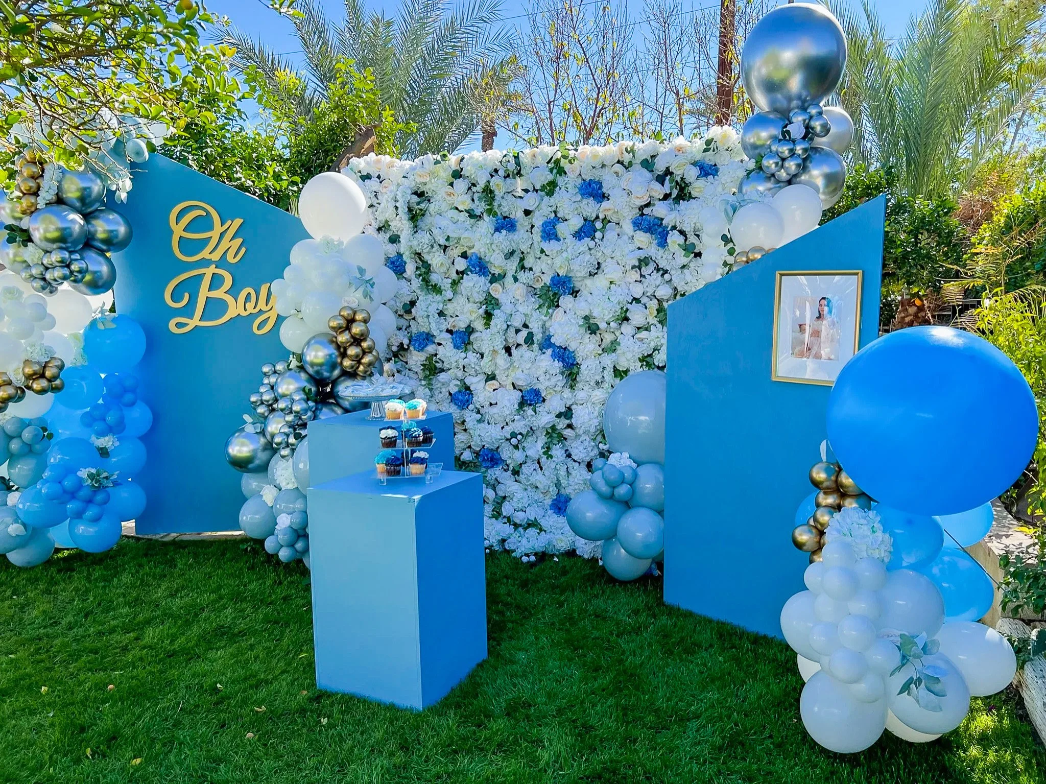 Outdoor baby shower decorations with a flower wall, balloons in white, blue, and metallic gold, and a framed photo of a woman in the foreground.