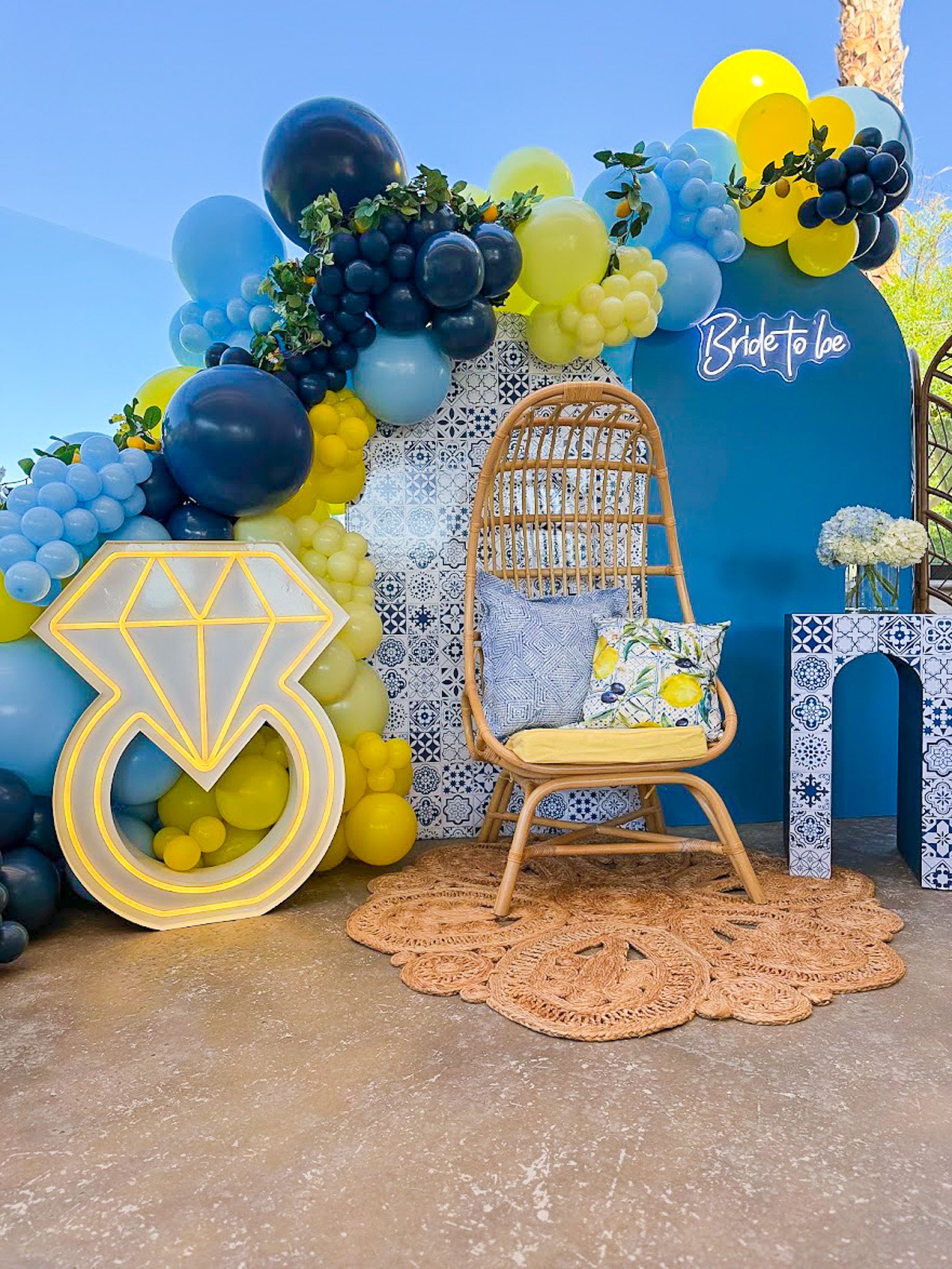 Decorative setup with blue, yellow, black, and clear balloons arranged around a blue backdrop with a neon sign that says 'Bridgerton'. A woven rattan chair with patterned cushions, a floral bouquet, and a glowing diamond-shaped light are also part of