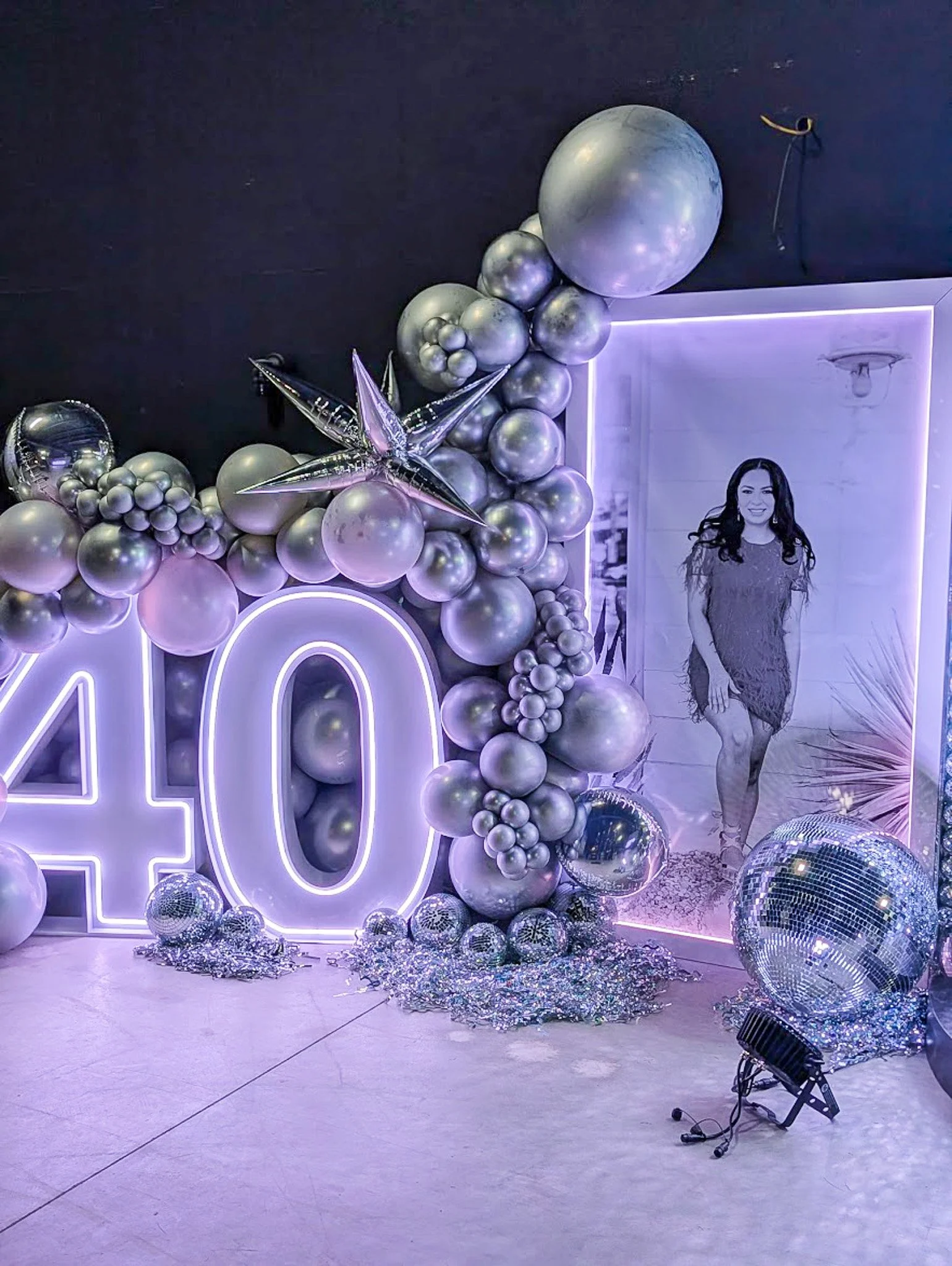 Wall decorated with silver and purple balloons, including a large number 20 outline with lights, on a party backdrop. There are large silver star-shaped balloons, disco balls, and confetti on the floor. A framed photo of a woman in a dress is on the 