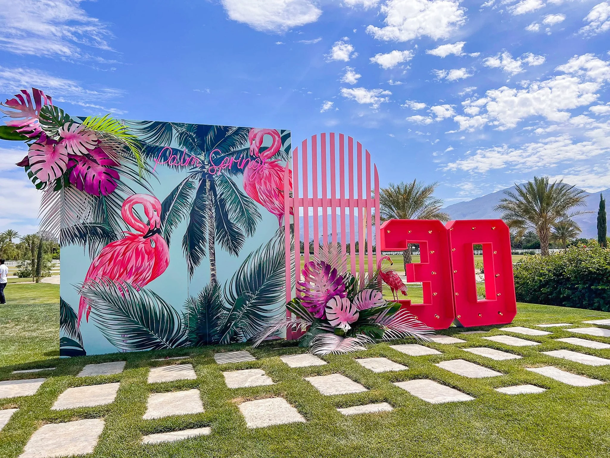 Colorful outdoor display with tropical-themed artwork and large pink block letter 'E' on green grass and blue sky background