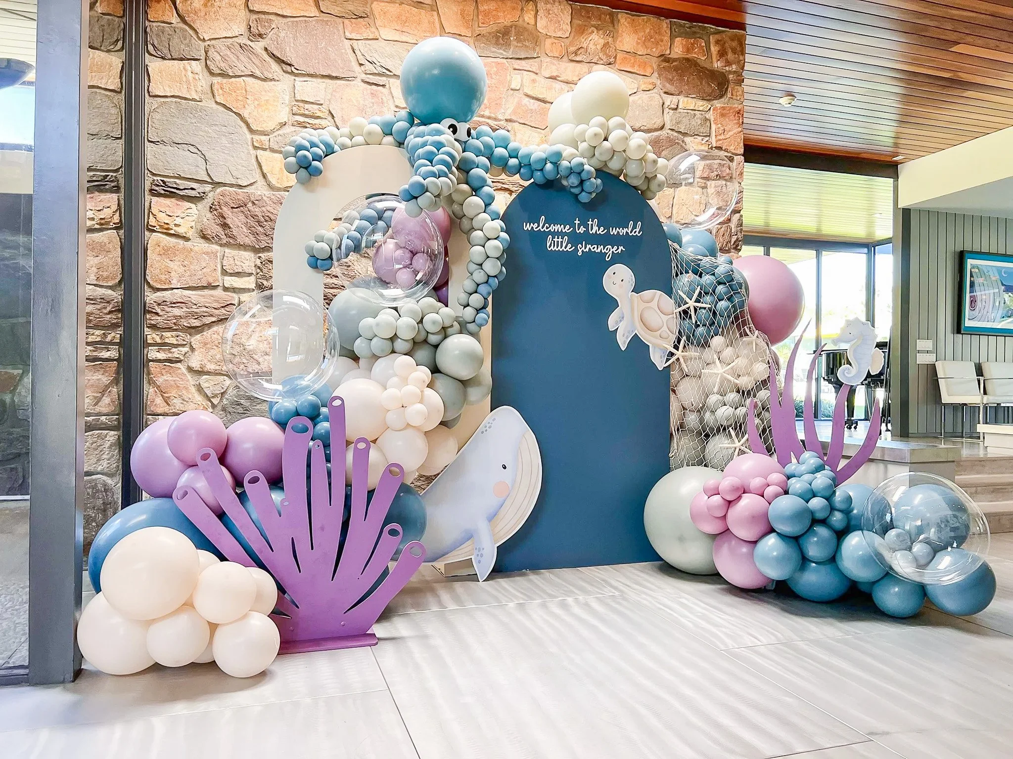 Underwater-themed balloon decoration featuring a turtle, seaplass, starfish, and sea plants in pastel colors of blue, pink, and white, with a sign reading 'welcome to the world little stranger'.