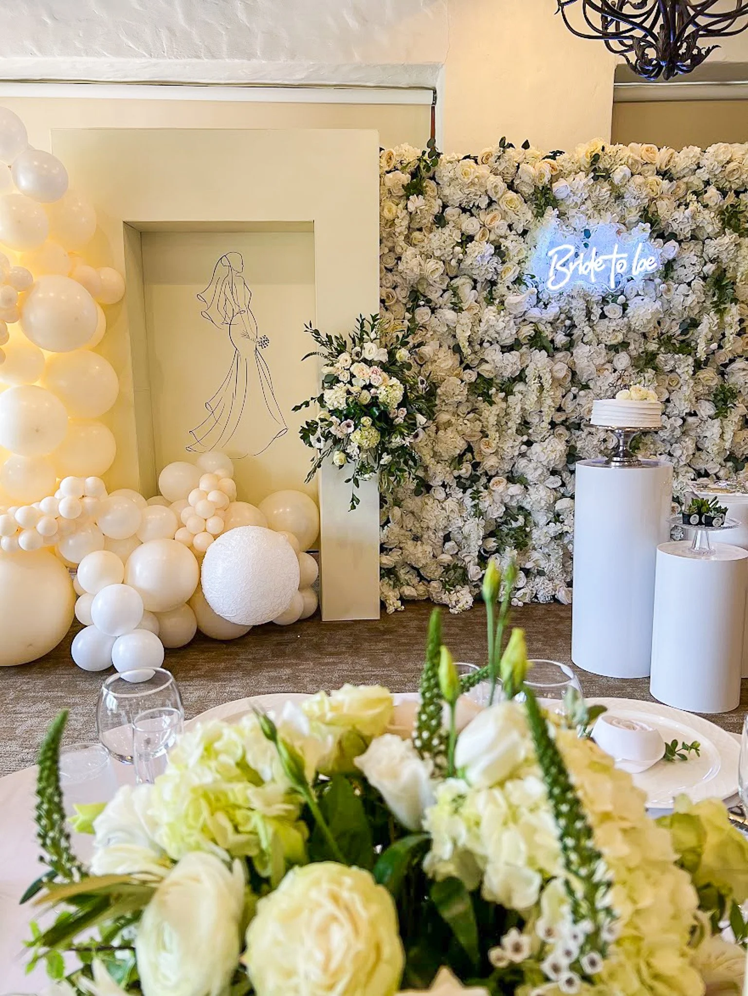 Wedding or event decor featuring a wall of white flowers with glowing neon sign that says "Bride to be," white and beige balloons, and a drawing of a bride in a long gown.
