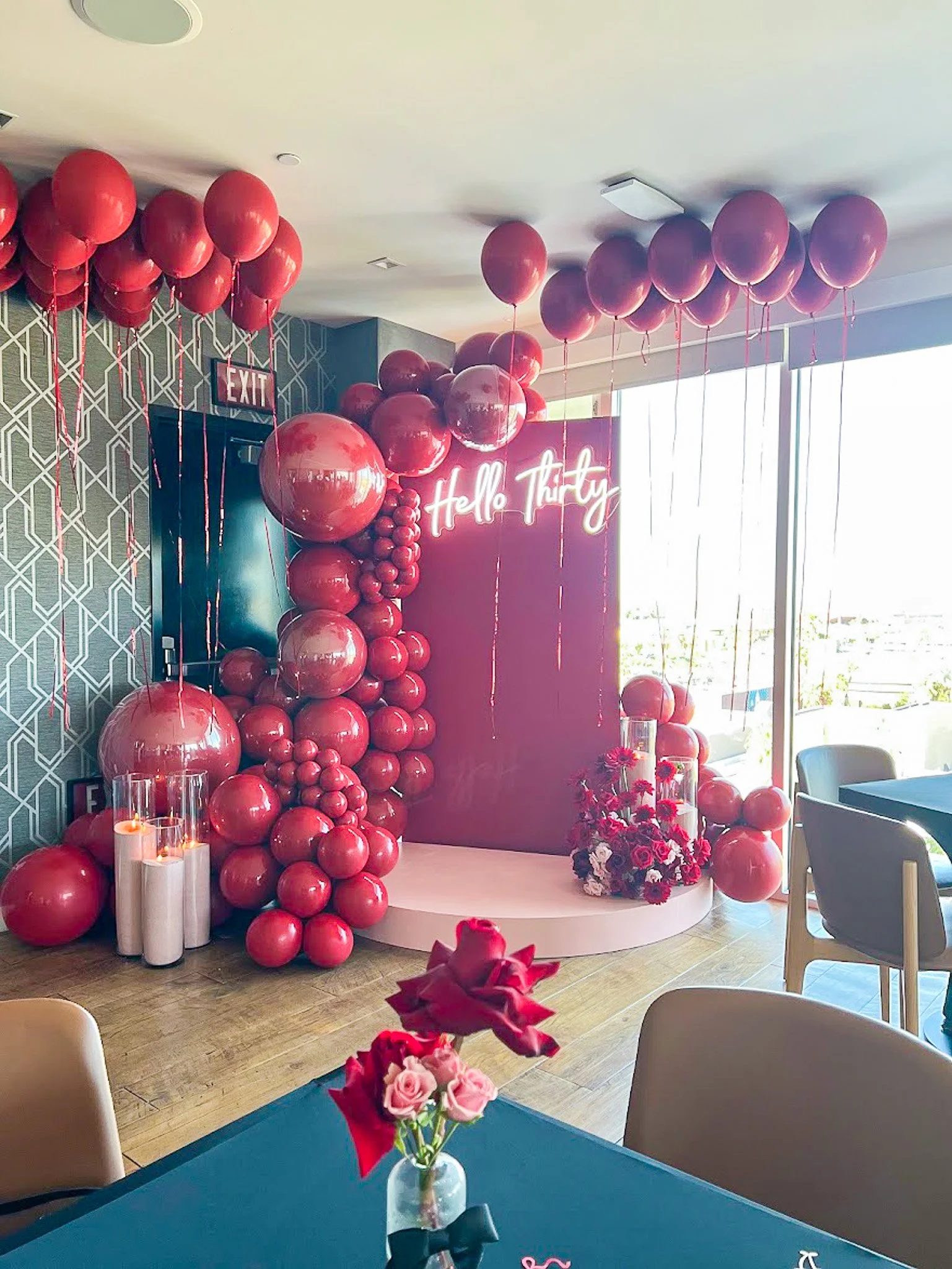 Celebration decorated room with pink and red balloons, a flower arrangement, and a neon sign reading 'Hello Thirty' for a 30th birthday.