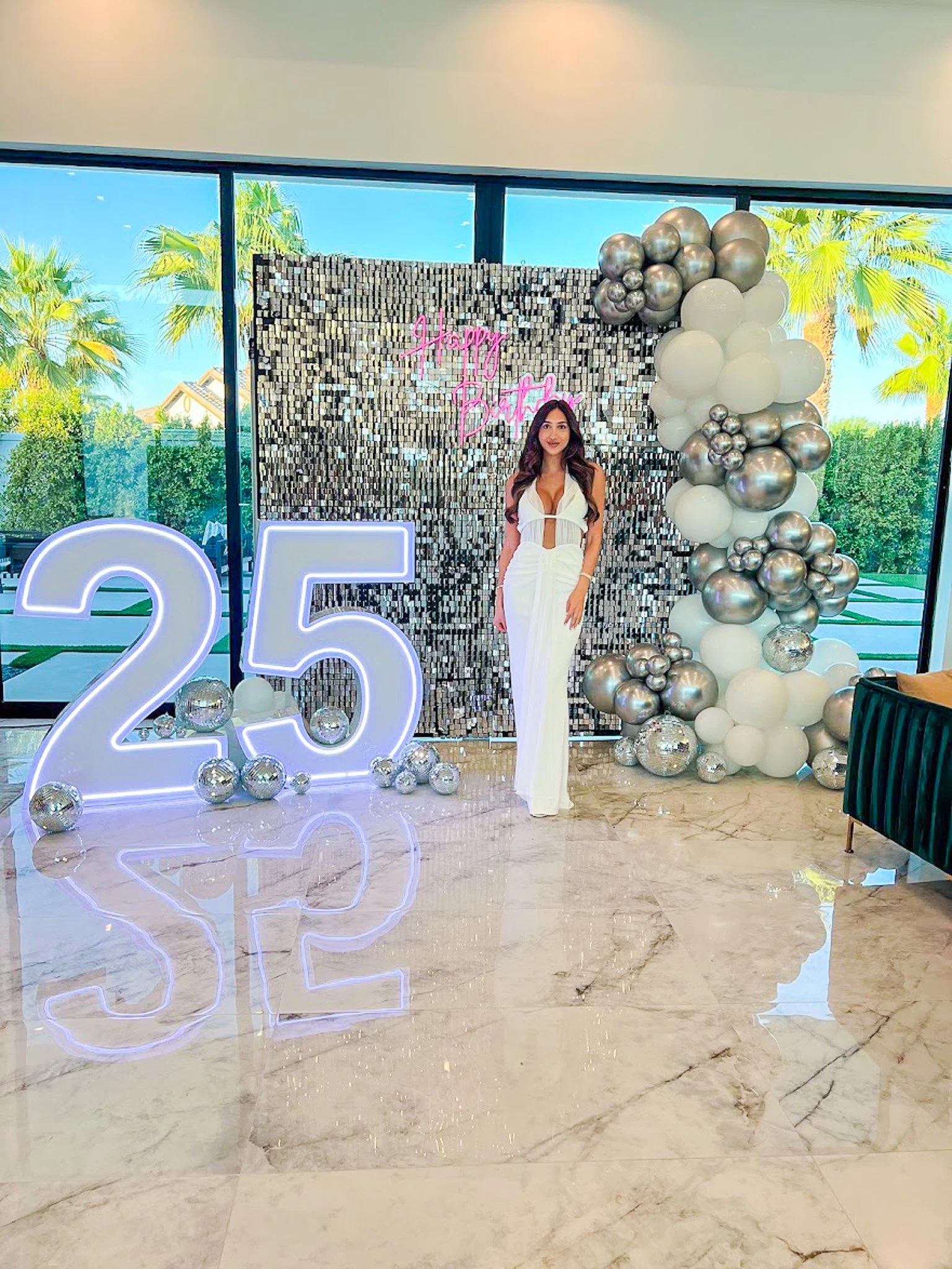 A woman in a white dress standing in front of a birthday backdrop decorated with balloons, a neon sign, and the number 25, with a large window showing trees outside.