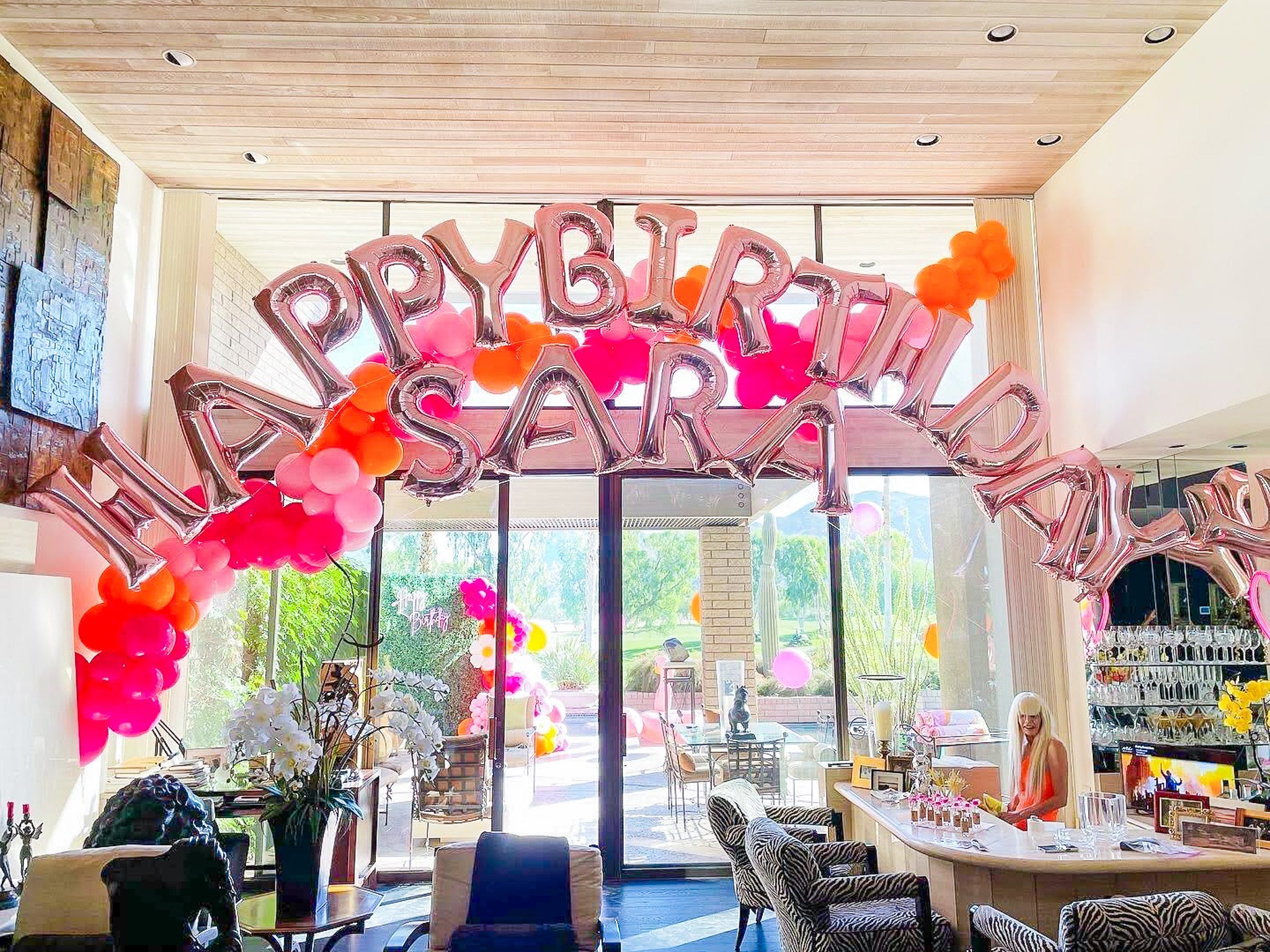 Celebration decorations with large pink and silver balloon letters spelling out 'HAPPY BIRTHDAY' surrounded by pink and orange balloons in a room with large windows