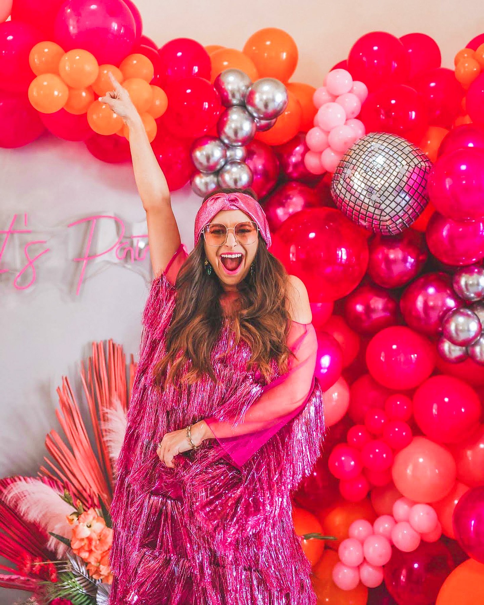 Woman smiling with excitement, wearing pink glasses and a pink headband, standing in front of a wall decorated with pink, red, orange, and silver balloons and some pink flowers.