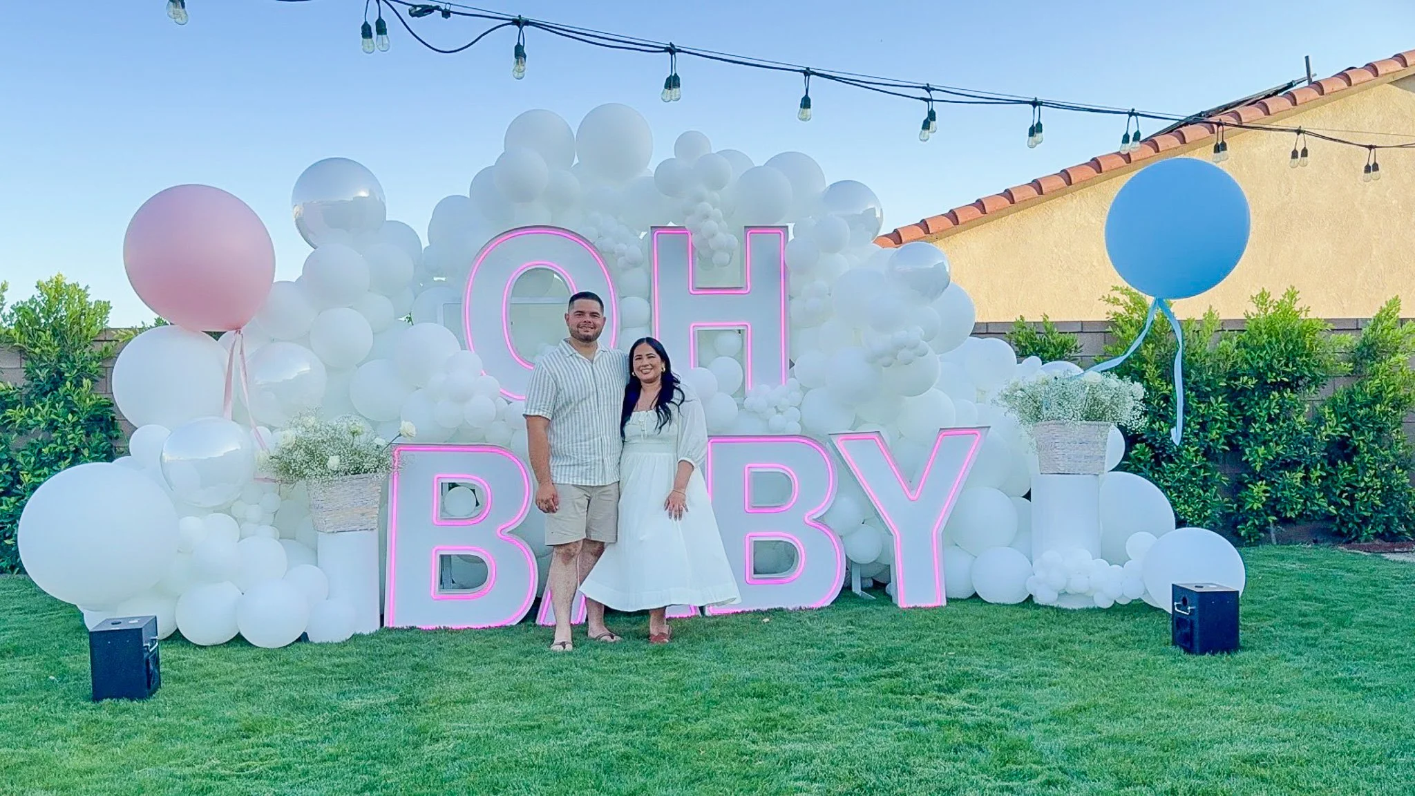 A couple standing in front of a decorated backdrop with balloons and neon pink letters spelling 'OH BABY'. The backdrop features white balloons and a neon sign, with string lights overhead, on a grassy outdoor area.