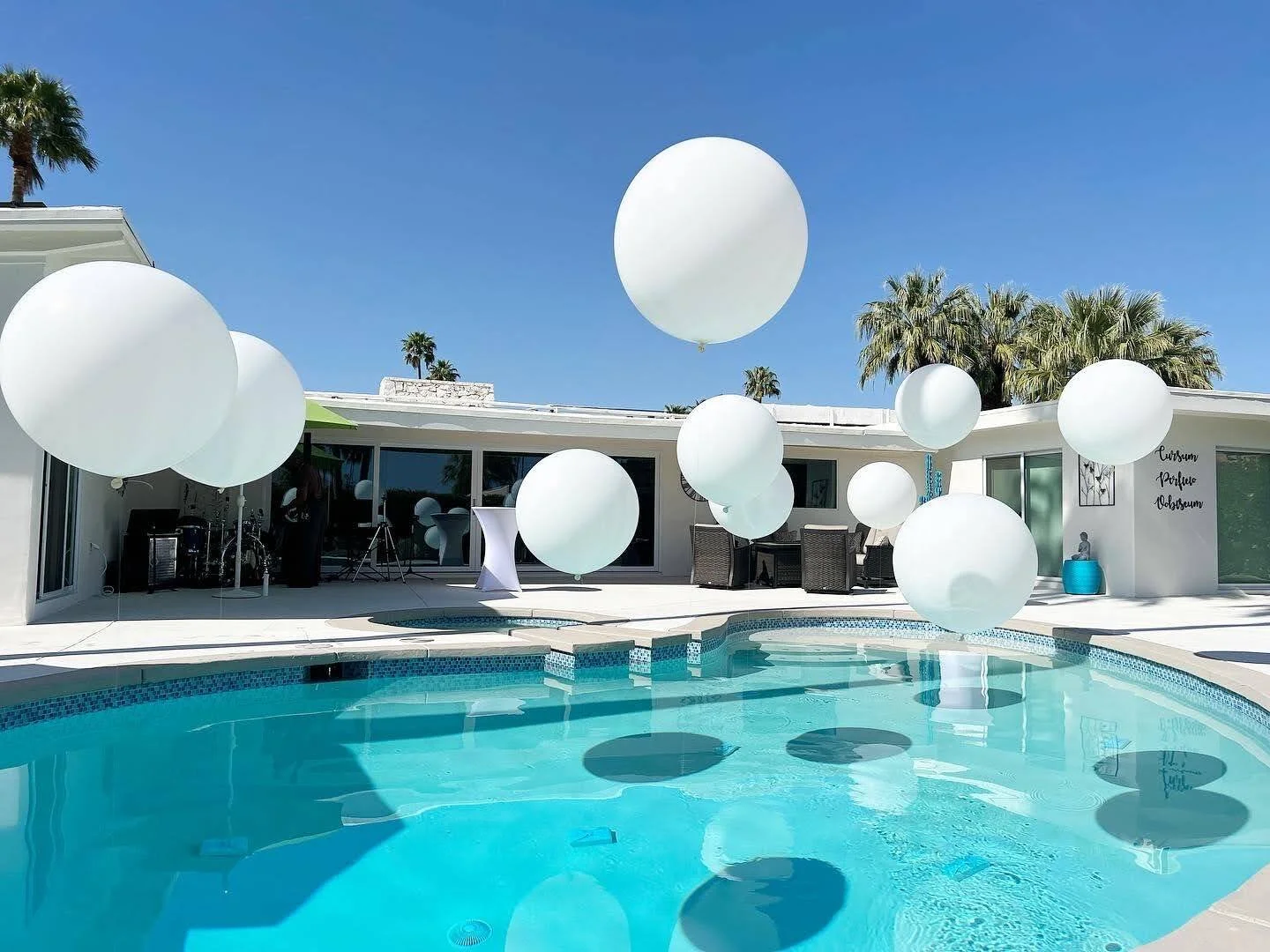 Poolside scene with large white balloons floating above the water and patio furniture, house with large windows, palm trees, and a clear blue sky.