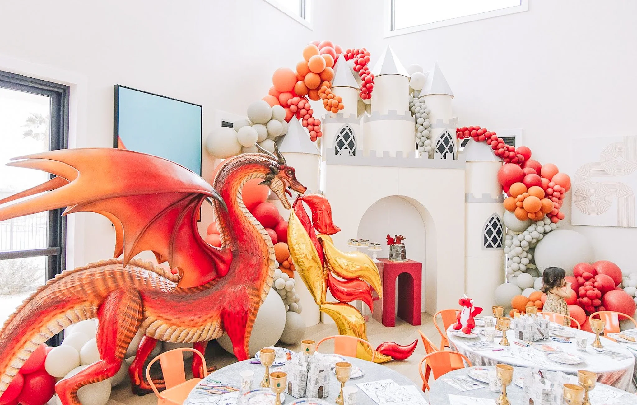 Party room decorated with a large birthday cake themed castle, orange and red balloons, fantasy dragon and unicorn inflatables, and a decorated table with party favors and seating.