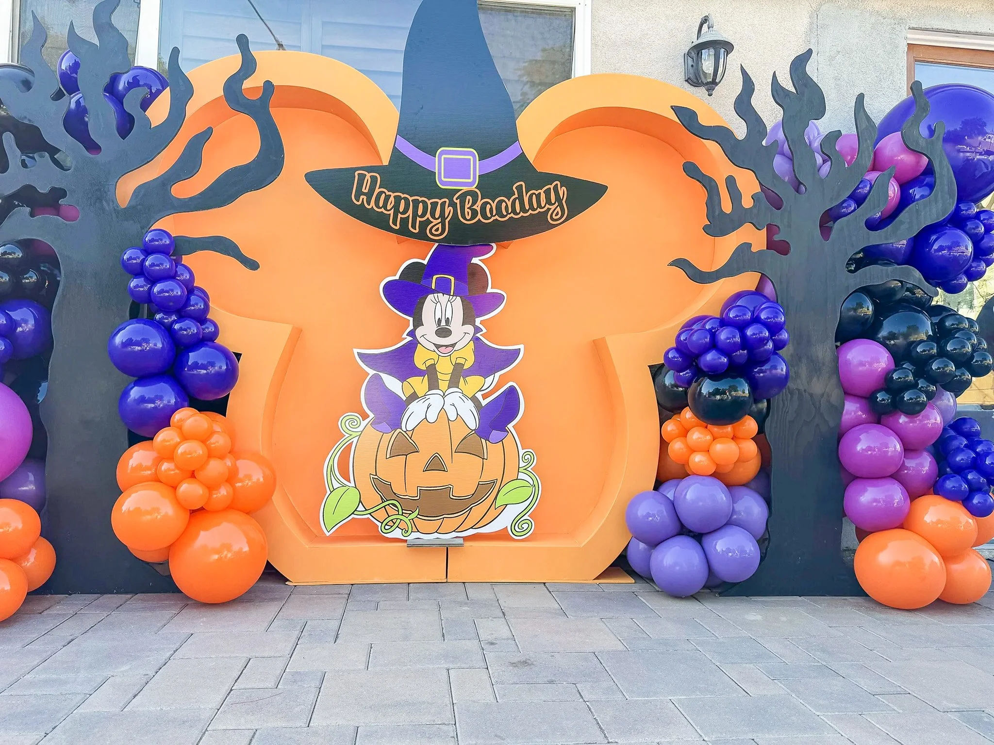 Outdoor Halloween decoration featuring a large orange pumpkin with a witch's hat and the words "Happy Badday" in the center. The decoration is surrounded by purple, orange, and black balloons arranged to resemble trees, with a house wall, window, and outdoor light in the background.