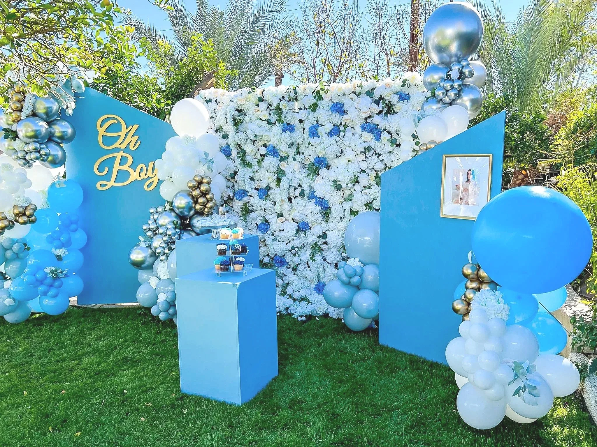 Decorative setup with a large floral display of white and blue flowers, blue boards with gold lettering, and various balloons in blue, white, and metallic colors, set outdoors on green grass with trees in the background.