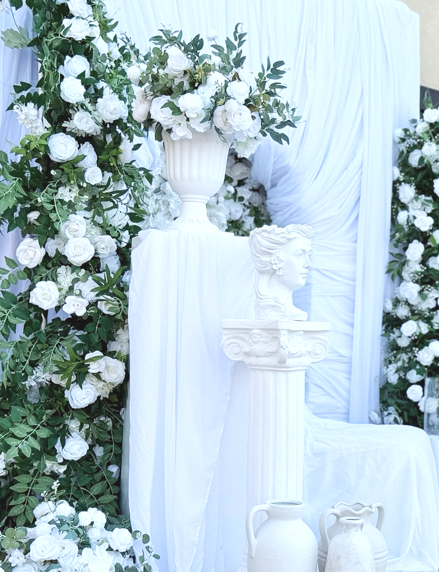 Decorative white floral display with white roses, a bust sculpture, and pottery vases against a white draped backdrop.