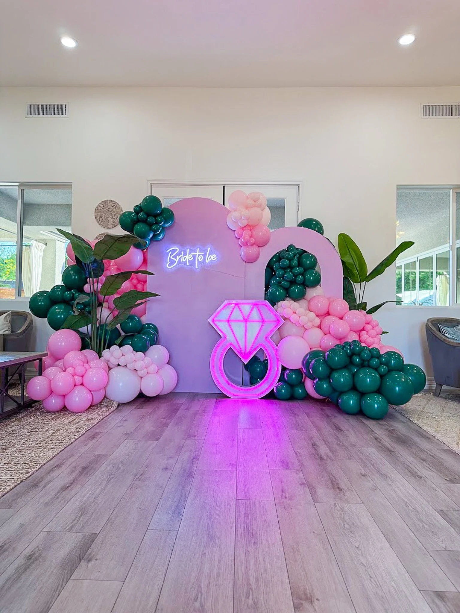 Decorative backdrop with pink and green balloons, a neon sign that says "Bride to be," and a neon diamond ring, set up for a bridal shower or bachelorette party in a bright indoor space.