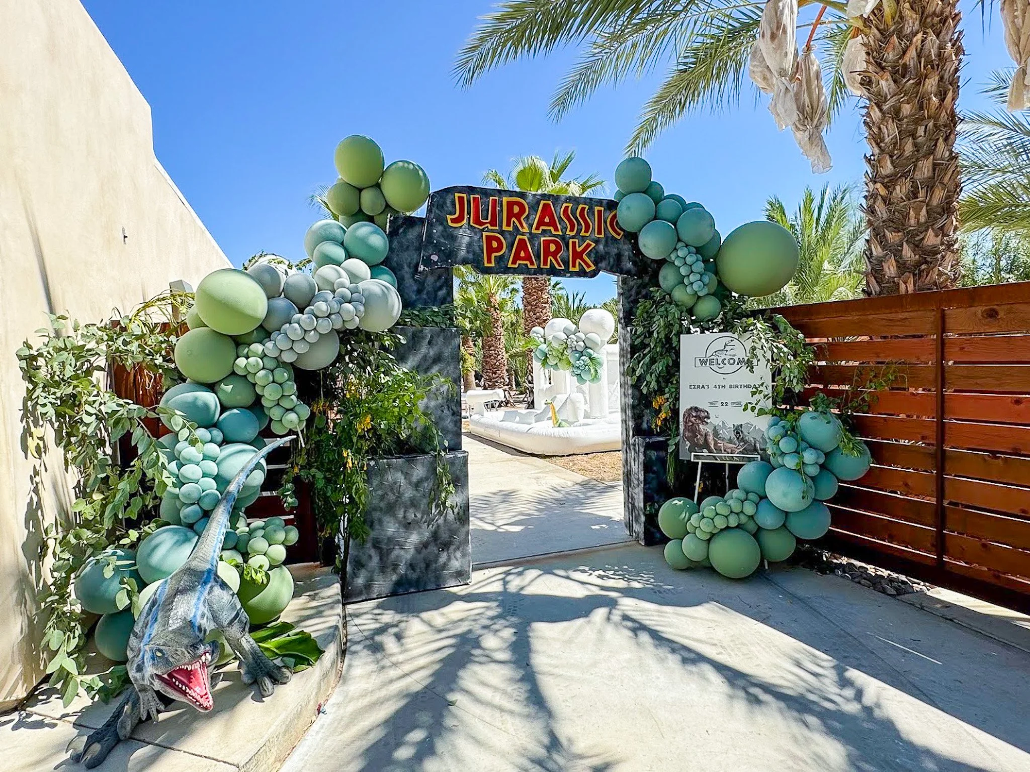 Decorations for Jurassic Park with green and white balloons framing the park entrance, including a dinosaur figure at the entrance and palm trees against a clear blue sky.