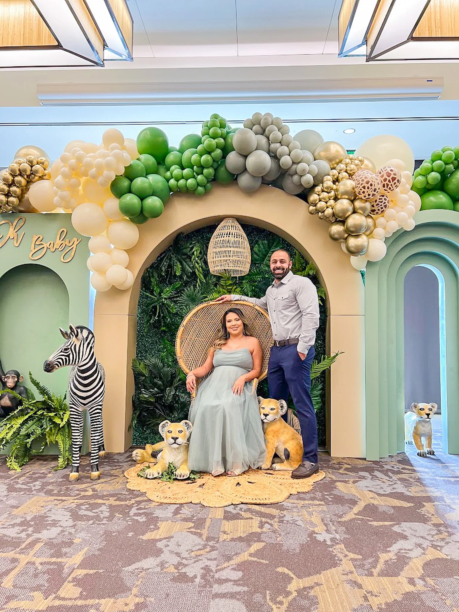 A couple at a safari-themed event, with a woman seated in a decorative chair and a man standing beside her. They are surrounded by jungle and animal decorations, including a zebra, lions, and additional tiger figures, with an arch of balloons in shad