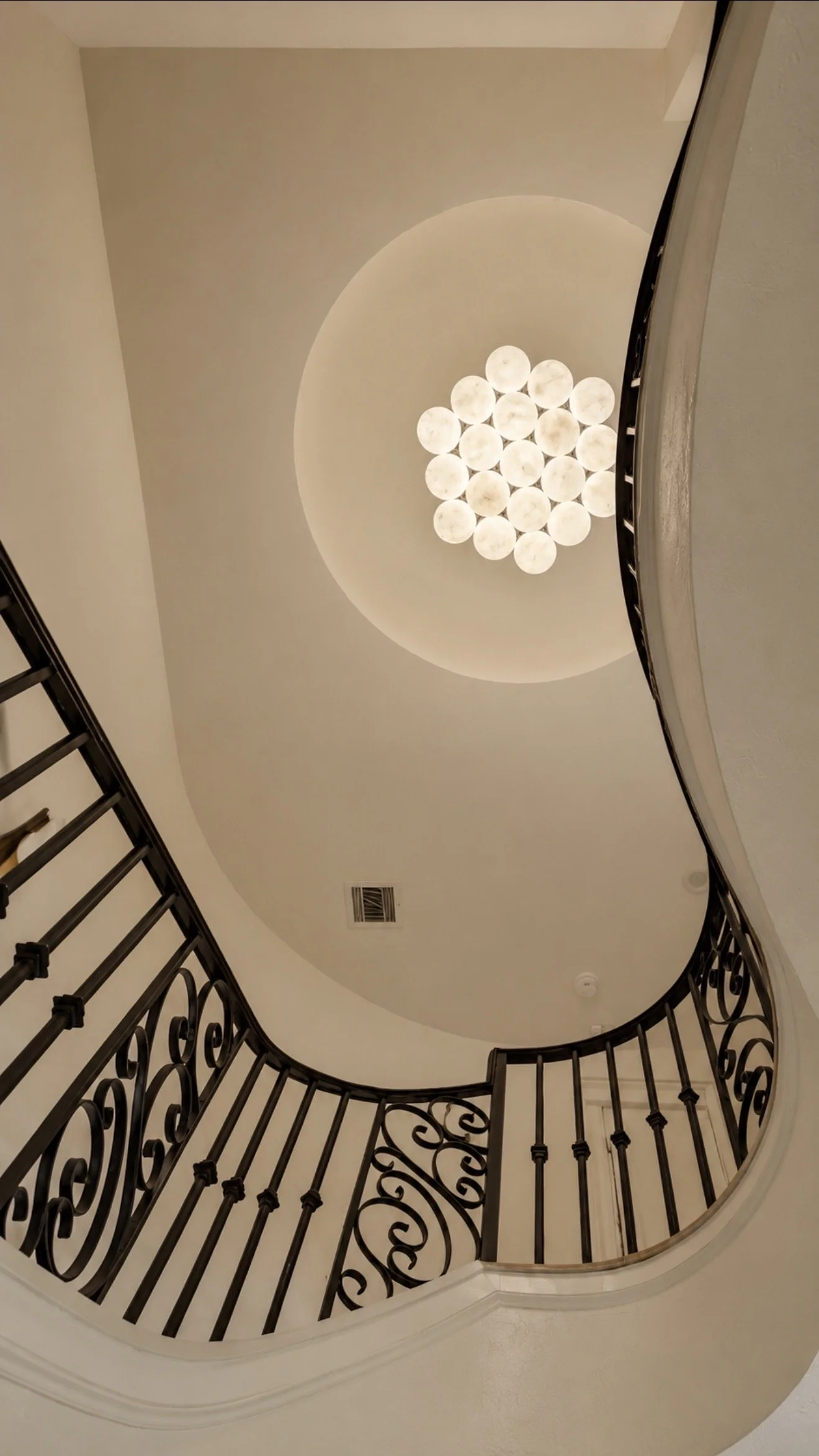 Stairwell viewed from above with a curved black iron railing, wood handrail, patterned flooring, and a ceiling light below.