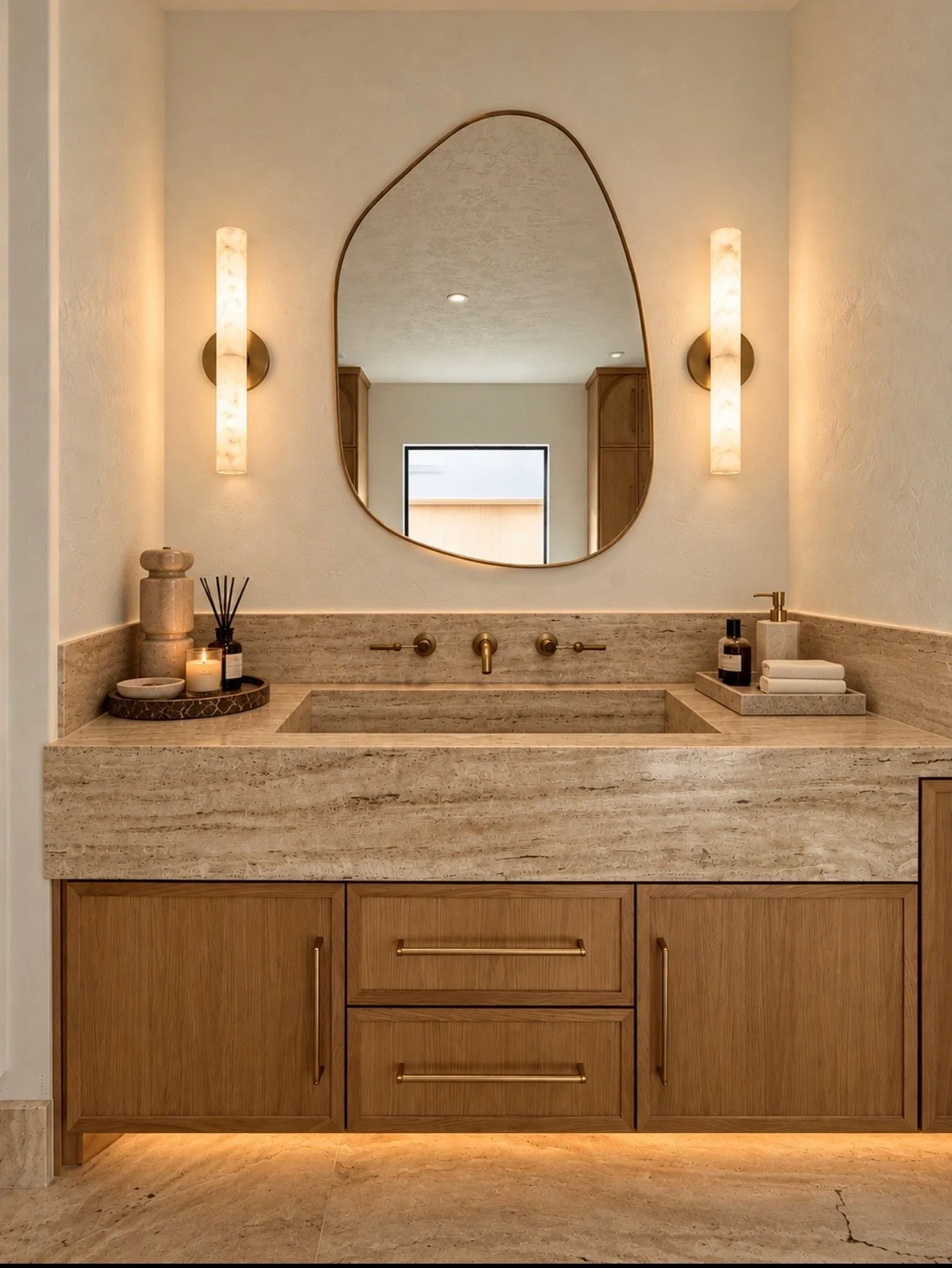 Warm natural stone bathroom vanity with integrated sink, arched mirror, wood cabinetry and wall-mounted brass fixtures.
