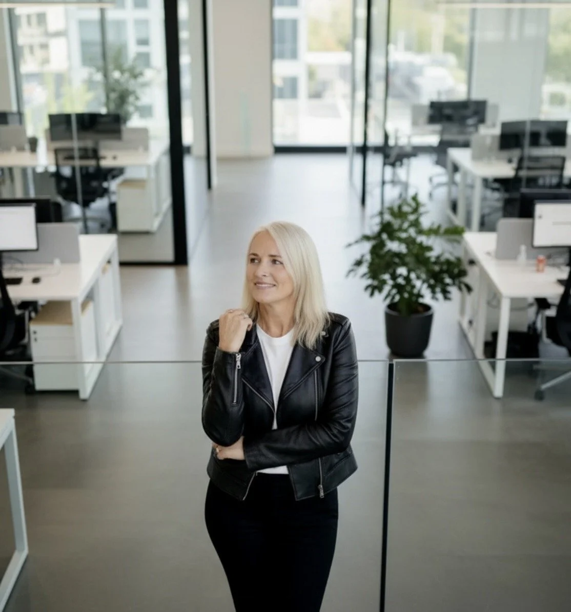 A woman with blonde hair, wearing a black leather jacket and white shirt, standing with her arms crossed and smiling in a modern open-concept office with multiple desks, chairs, and large windows letting in natural light.