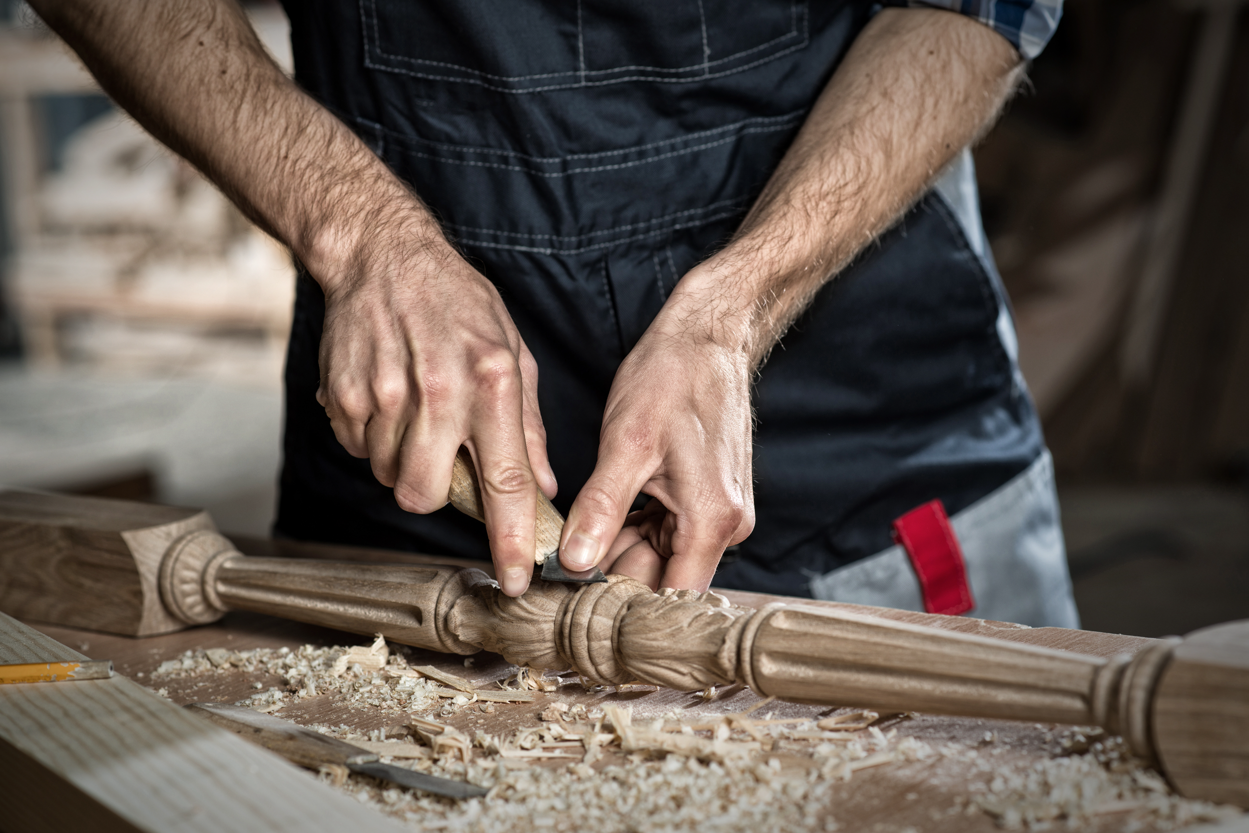 A craftsman wearing a black apron is carving a wooden piece with a chisel and mallet in a woodworking shop.