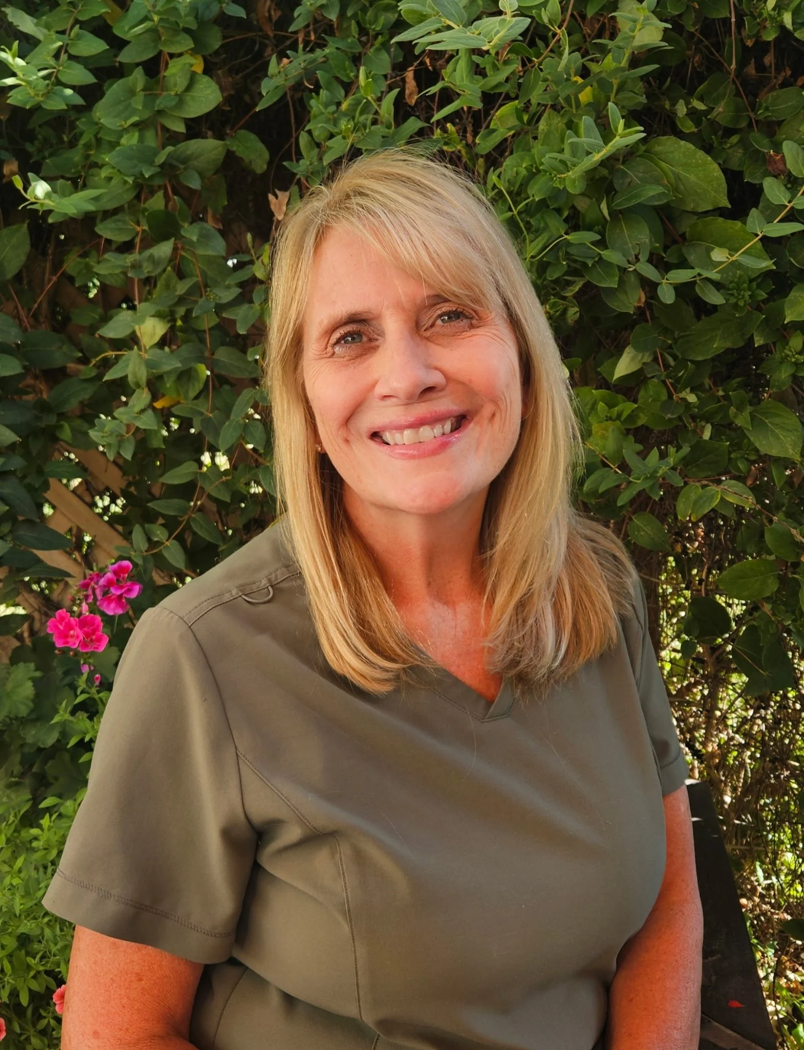 A woman with blonde hair smiling outdoors in front of a green leafy hedge with pink flowers.