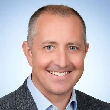 Headshot of a smiling man with short hair, wearing a suit jacket and light blue shirt, against a light blue background.