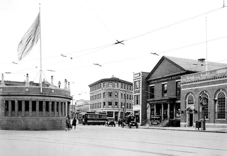 Historical street scene in black and white featuring a corner building with signs for Harvard Cooperative Society, a streetcar, and pedestrians, with a large flag and street wires overhead.