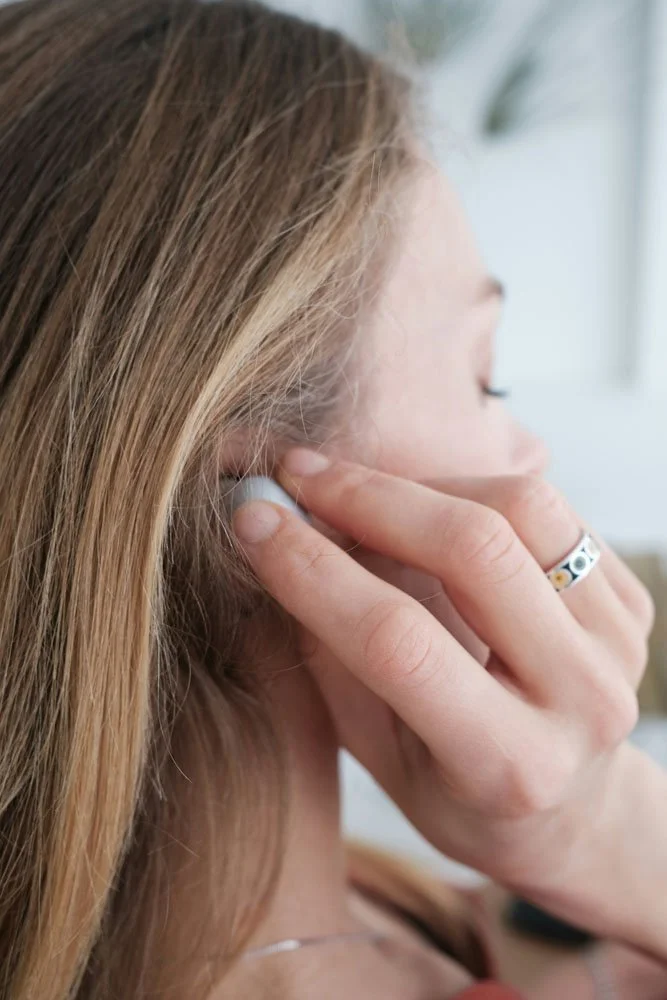 Close-up of a woman with blonde hair placing an earbud in her ear, wearing a silver ring with colorful designs on her finger.
