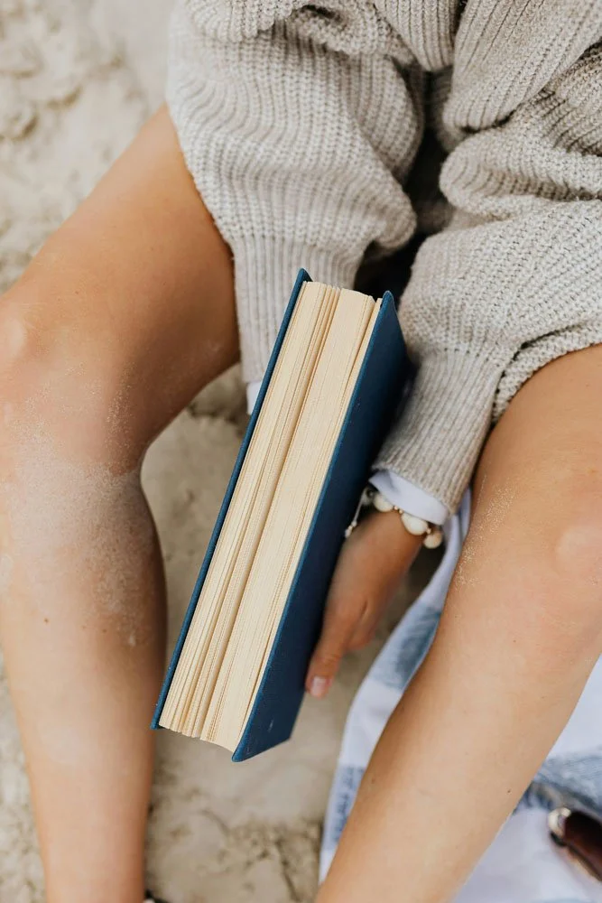 A person sitting outdoors on sandy ground, holding a closed hardcover blue book with visible cream-colored pages, wearing a beige knit sweater, white jewelry, and sand on their arms and legs.