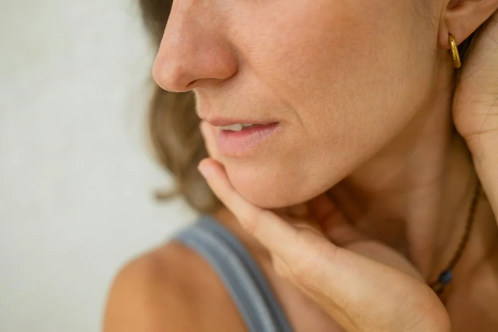 Close-up of a woman with light skin and earrings, touching her chin with her hand, wearing a tank top.