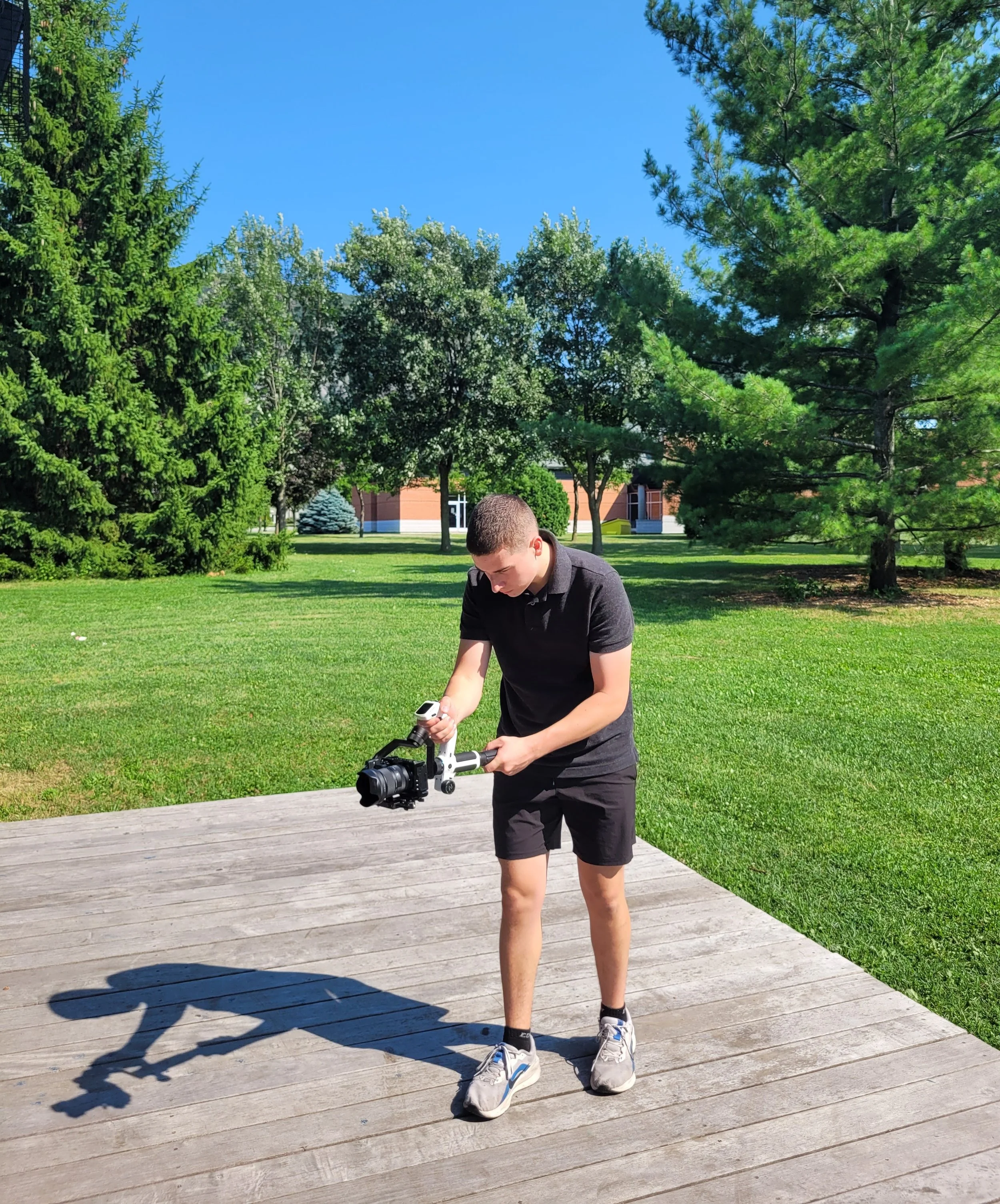 Un jeune homme marchant sur une plateforme en bois dans un parc, manipulant une caméra ou un drone, sous un ciel bleu clair entouré d'arbres verts.
