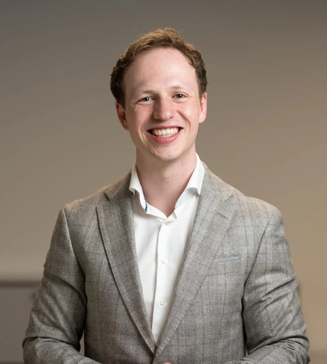 A young man with curly brown hair, smiling, wearing a light gray blazer and white dress shirt, against a plain neutral background.