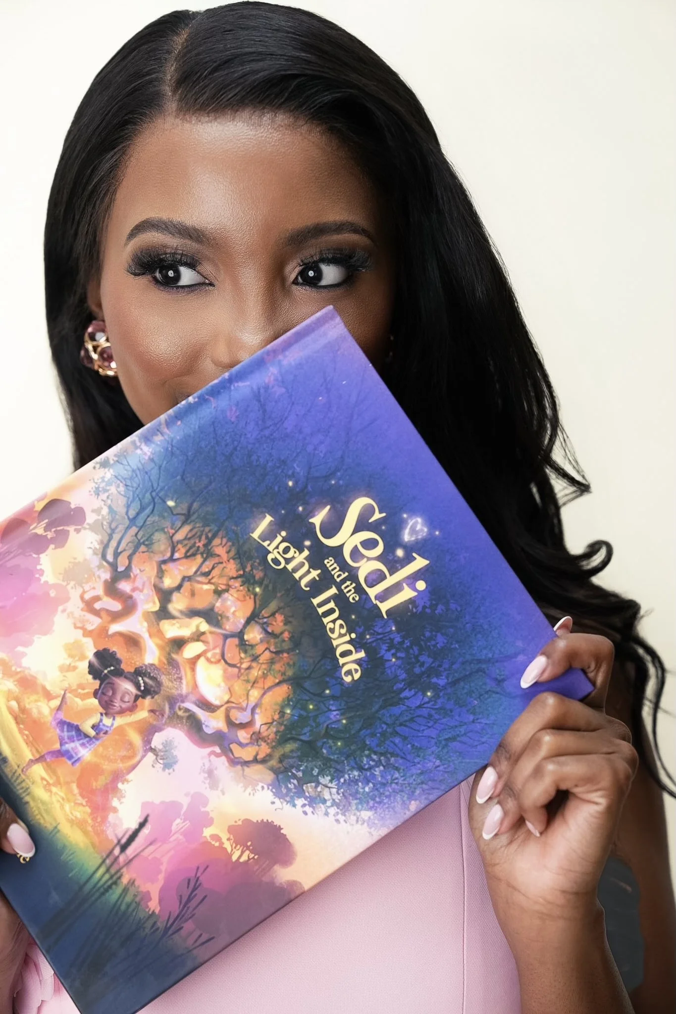 A woman with dark hair, makeup, and earrings is holding a colorful book titled 'Jedi and the Light Inside,' partially covering her face. She is wearing a pink top and has neatly manicured nails.