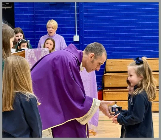 A priest in purple robes is giving an award or medal to a young girl with a bow in her hair during a school assembly or ceremony, with other children and an adult woman observing.