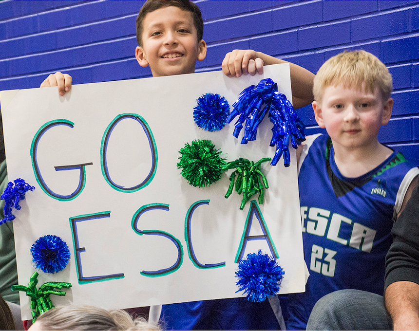 Two boys holding a sign that says "GO ESC" decorated with blue and green pom-poms, with a blue brick wall in the background.
