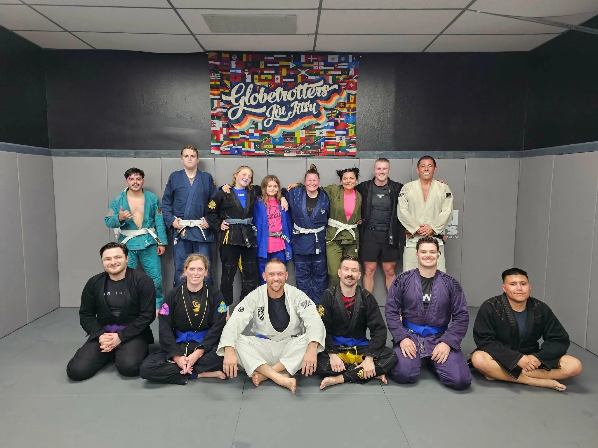 Group of people in martial arts gis and uniforms, some sitting and some standing, in a dojo with a colorful flags banner and the words 'Globetrotters Jai Jitsu' on the wall behind them.