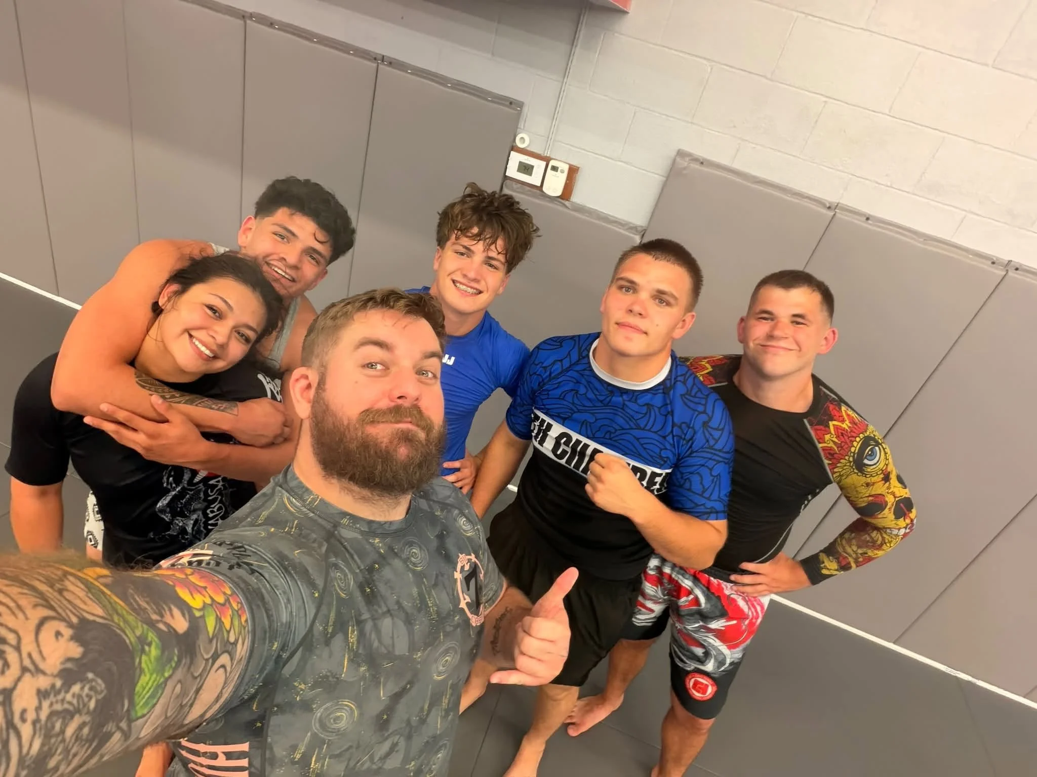 A group of five young adults posing for a selfie in a sports facility, with some showing thumb-up gestures. Four of them are men, and one is a woman, all dressed in casual and athletic clothing.