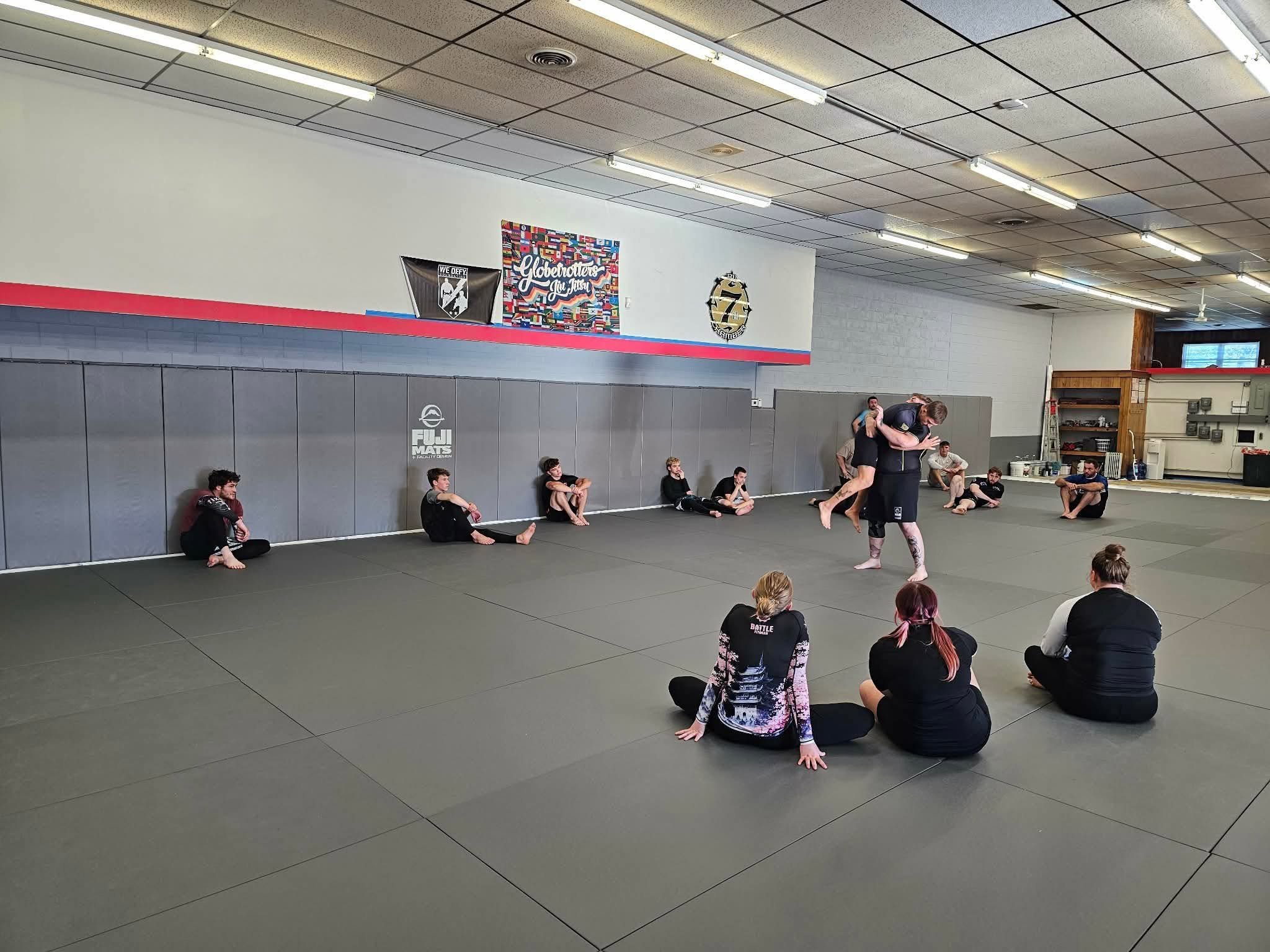 Martial arts class with students sitting on mats against a wall, a man demonstrating technique with a partner, and posters on the wall, including one that says "Globetrotters Jiu Jitsu".