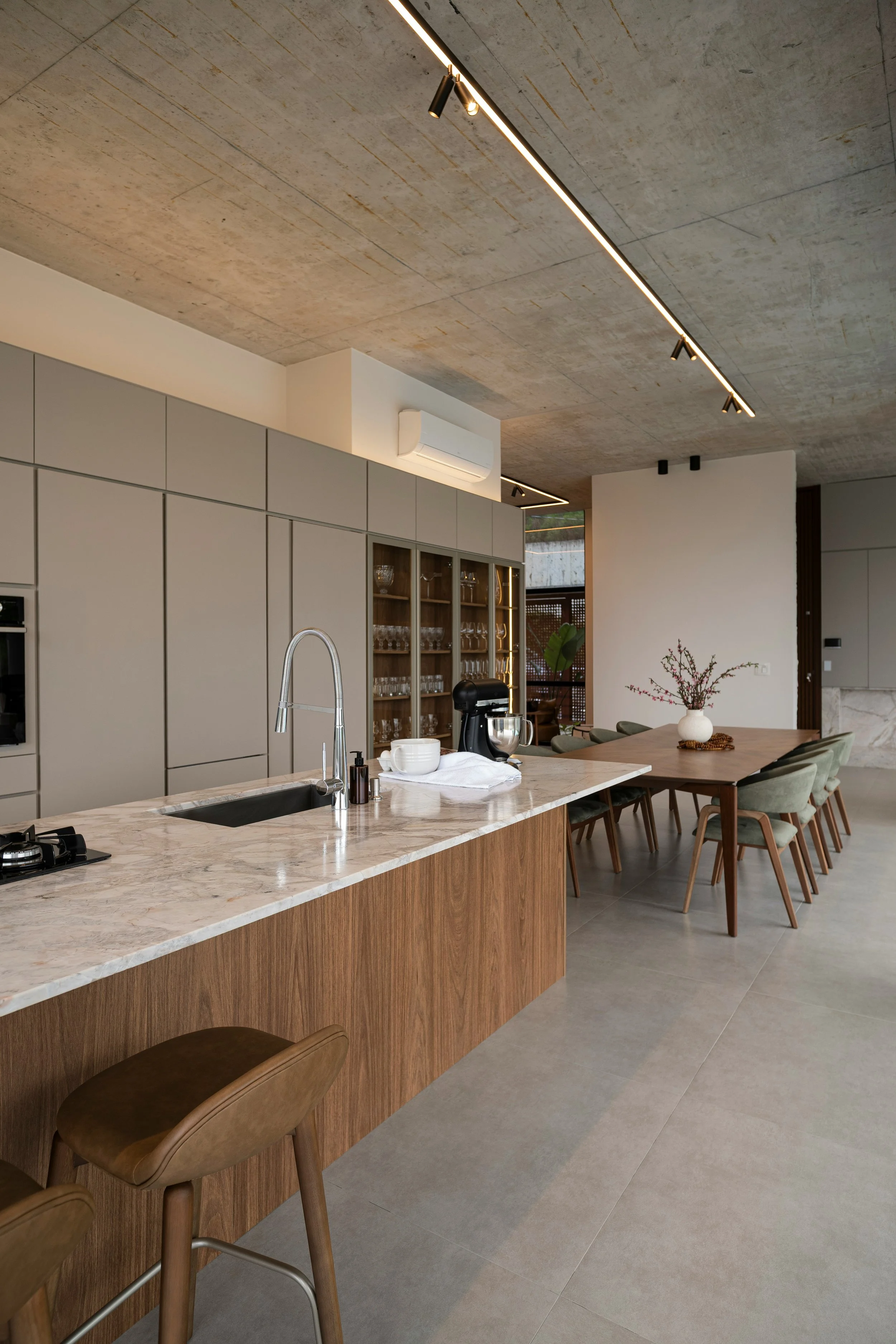 Modern kitchen with a marble island counter, light gray cabinetry, a built-in oven, and a dining area with a wooden table and green chairs. The ceiling has minimalist track lighting, and there are decorative flowers on the table.