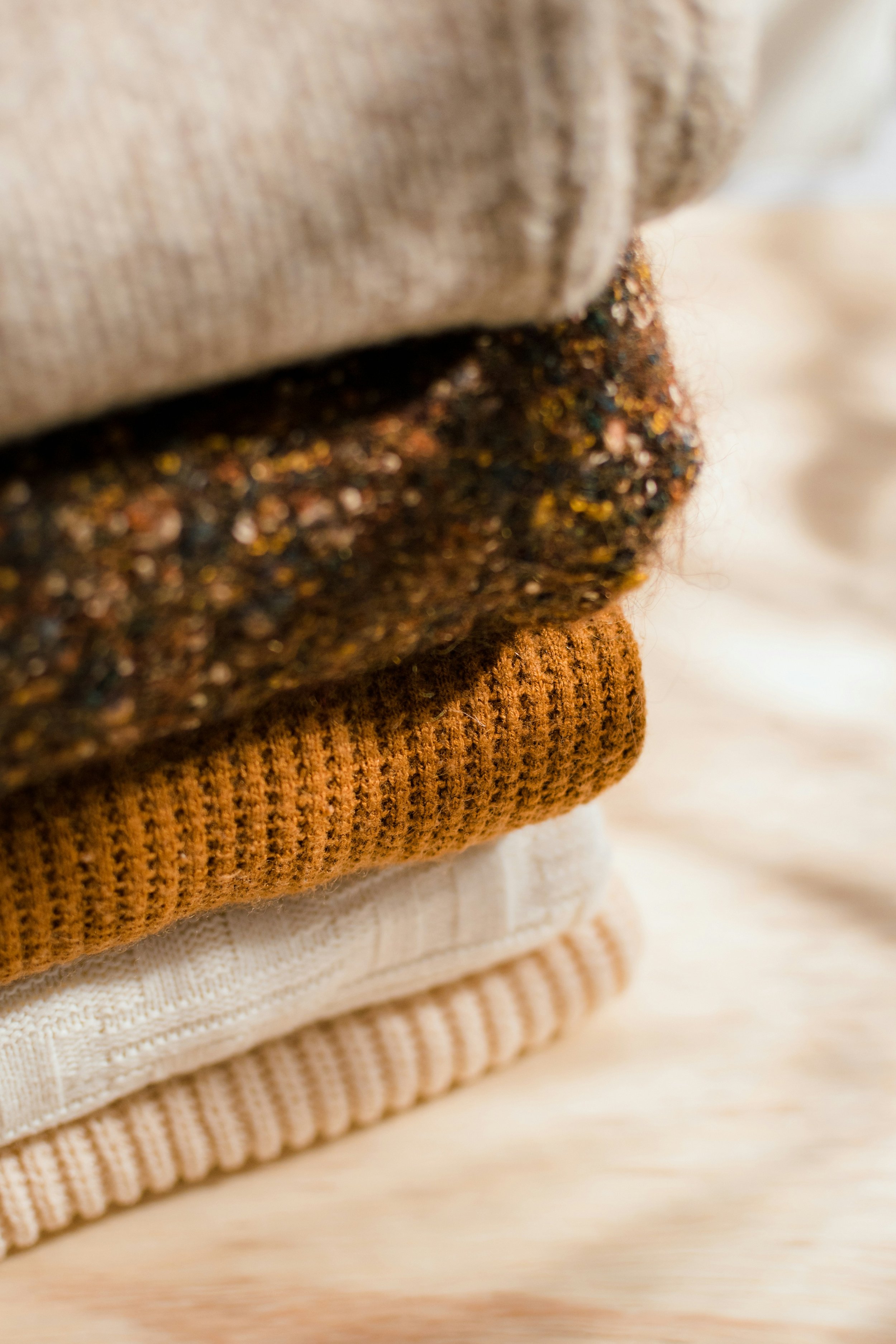 Stacked folded sweaters in neutral and warm tones, including beige, brown, and cream, on a light wooden surface.