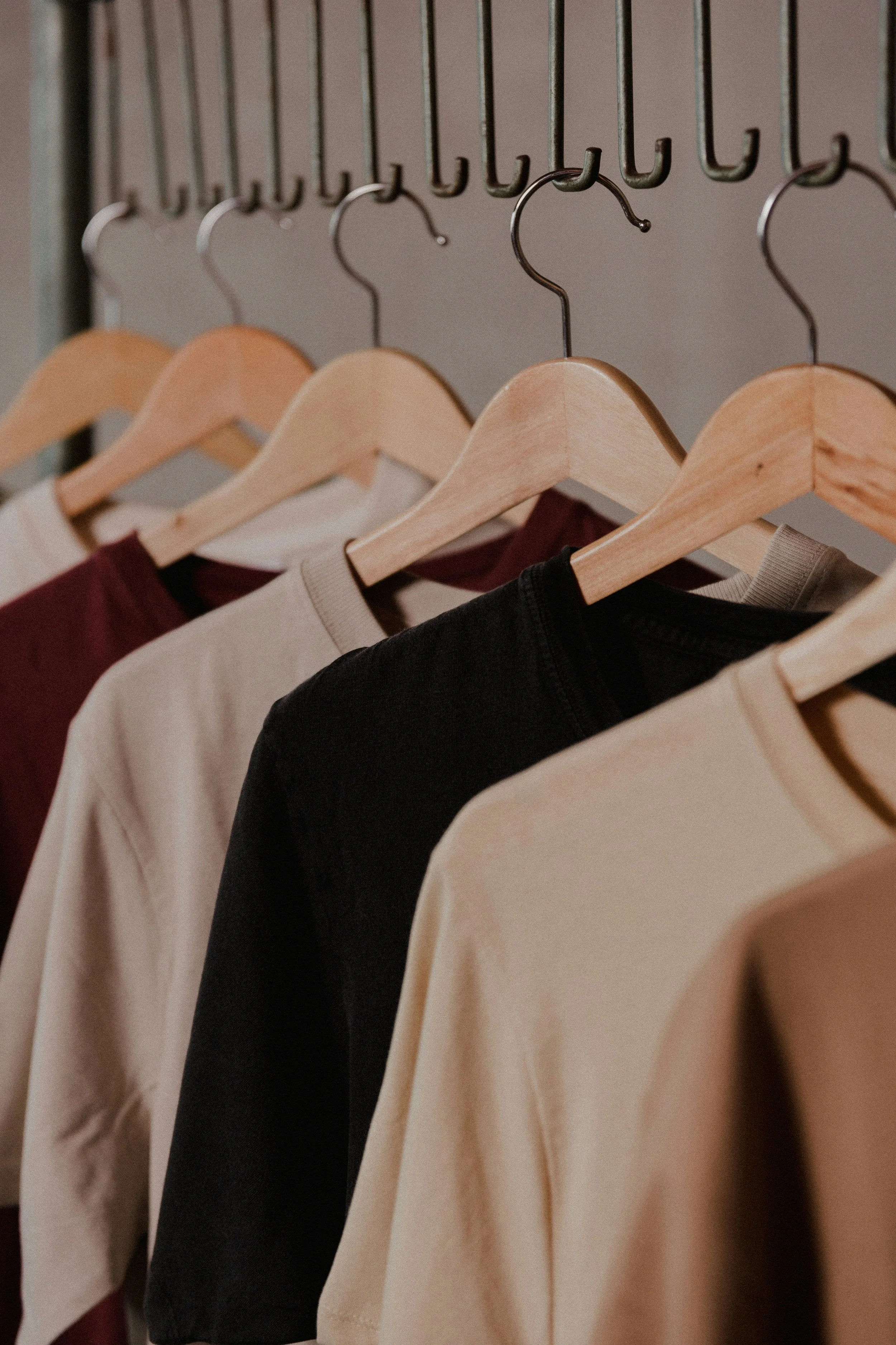 Clothing items hanging on wooden and metal hangers on a rack, including beige, black, and maroon shirts.